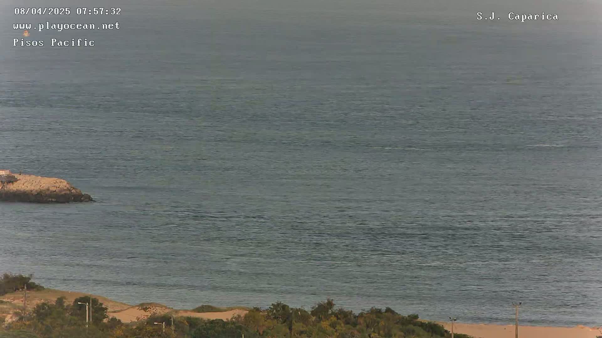 A rocky outcrop juts into a calm, gray-green ocean under an overcast sky, with vegetation and a sandy beach visible in the foreground.
