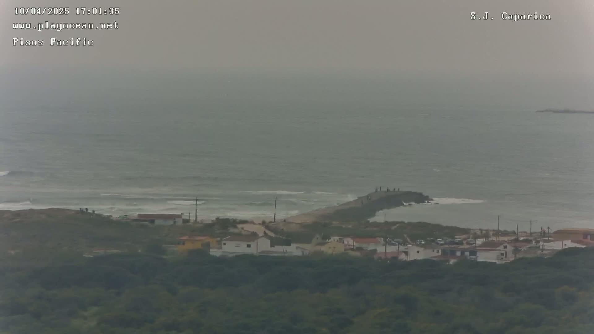 A hazy, overcast day shows a rocky coastline with a small pier, a cluster of buildings, and a swath of dark green vegetation.