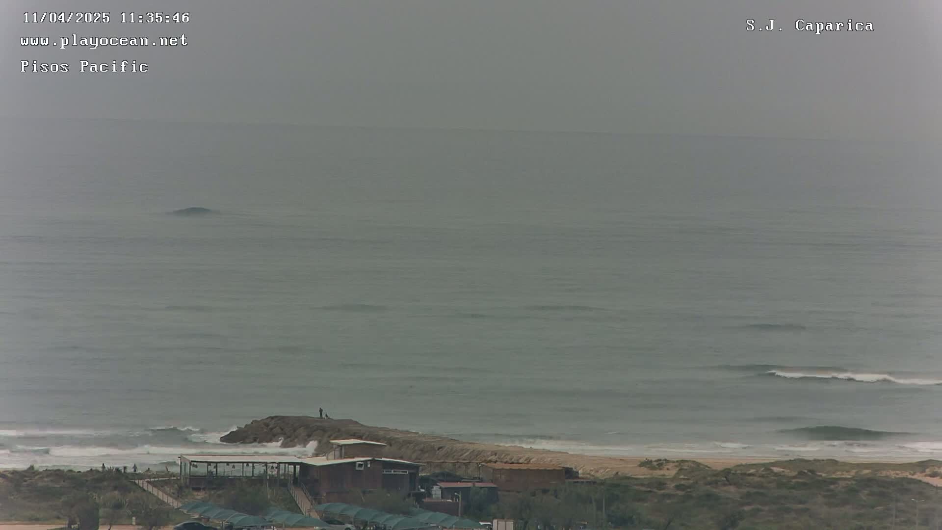 A hazy overcast day reveals a beach with small waves rolling onto the shore, near a structure with several buildings and a person standing on a rock jetty.