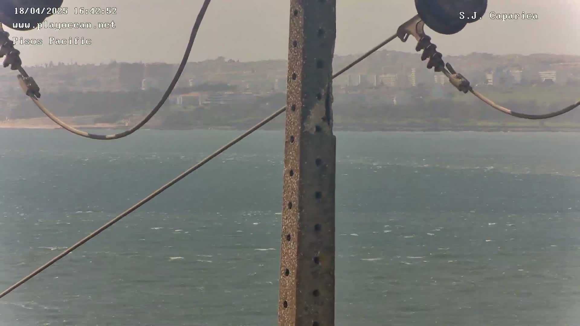 A weathered pole stands in the foreground against a slightly hazy background of a wavy ocean and distant buildings under a somewhat overcast sky.