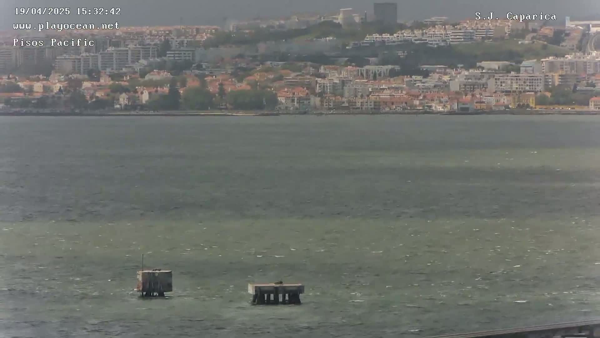 A coastal city is visible across a choppy body of water, with two small structures in the foreground.