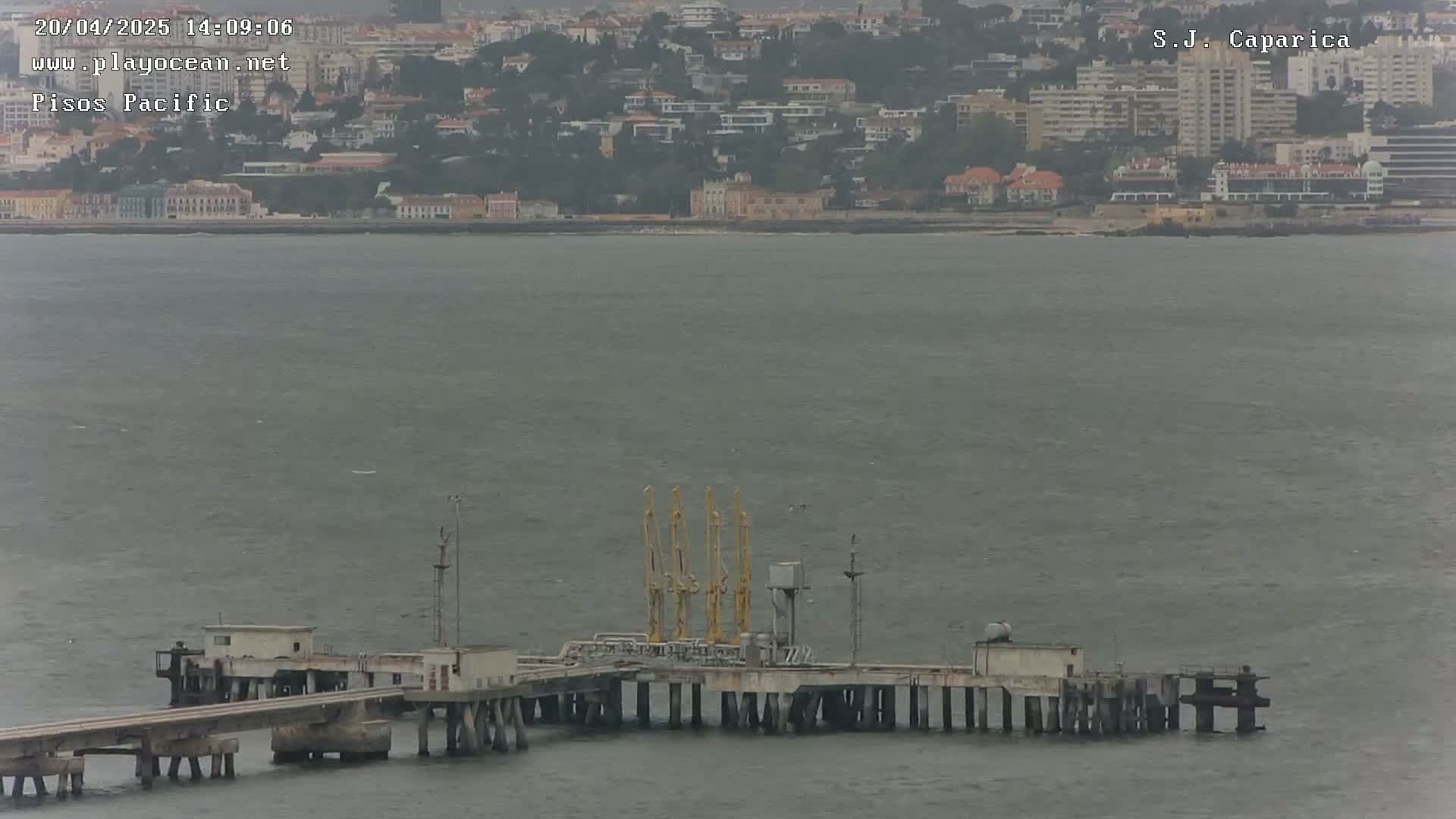 A long pier with several small buildings and yellow cranes extends into a calm, gray body of water, with a distant cityscape visible in the background under an overcast sky.