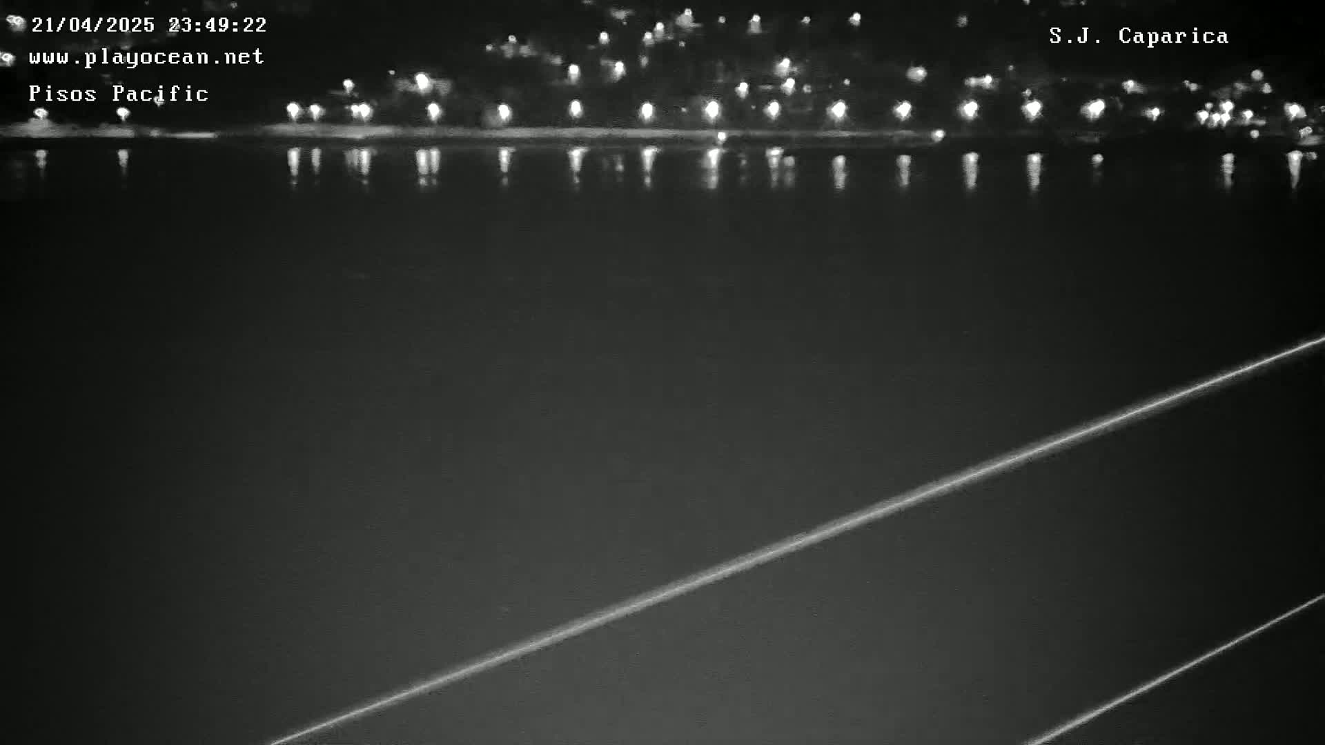 A nighttime view shows a dark body of water reflecting the lights of a distant town under a clear sky.