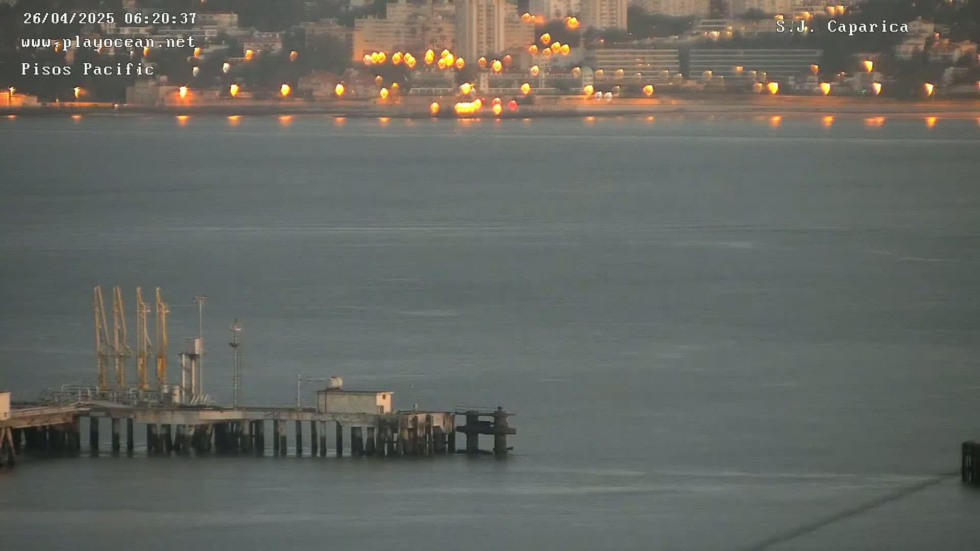 A pier with several cranes extends into a calm, gray body of water in front of a city skyline at sunset.