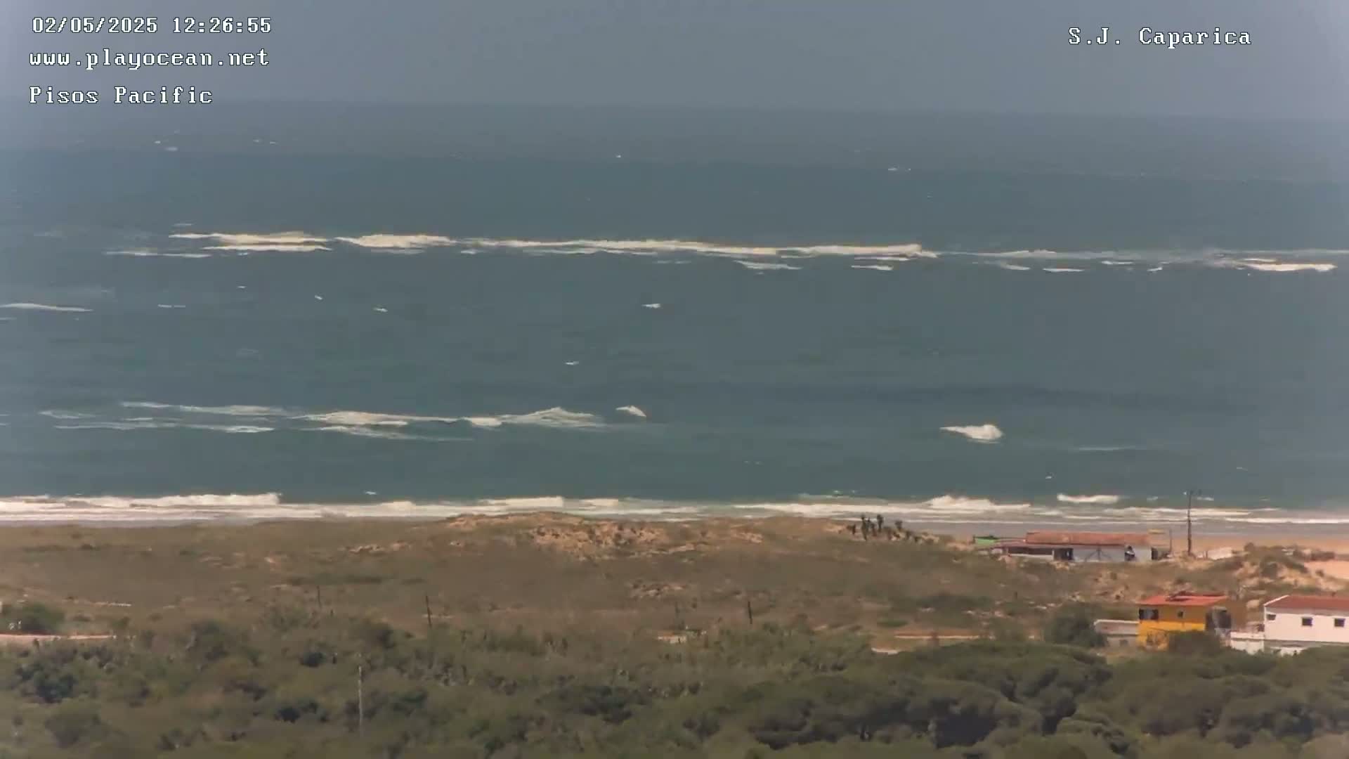 A coastal view shows a somewhat choppy ocean with small waves breaking on a sandy beach, beyond which is tan-colored scrubland, some small buildings, and dark green vegetation.