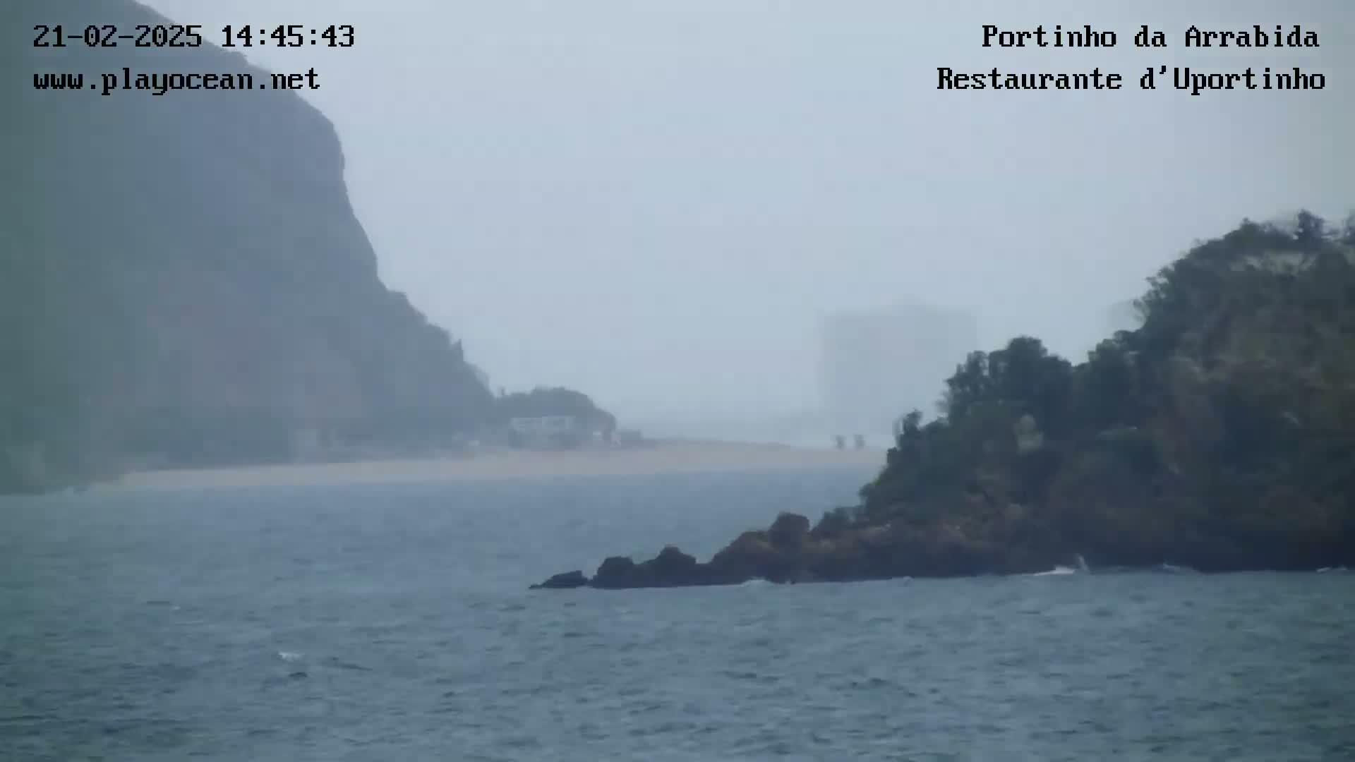 A hazy, overcast day shows a rocky shoreline with a small island and a sandy beach visible in the distance across a choppy sea.