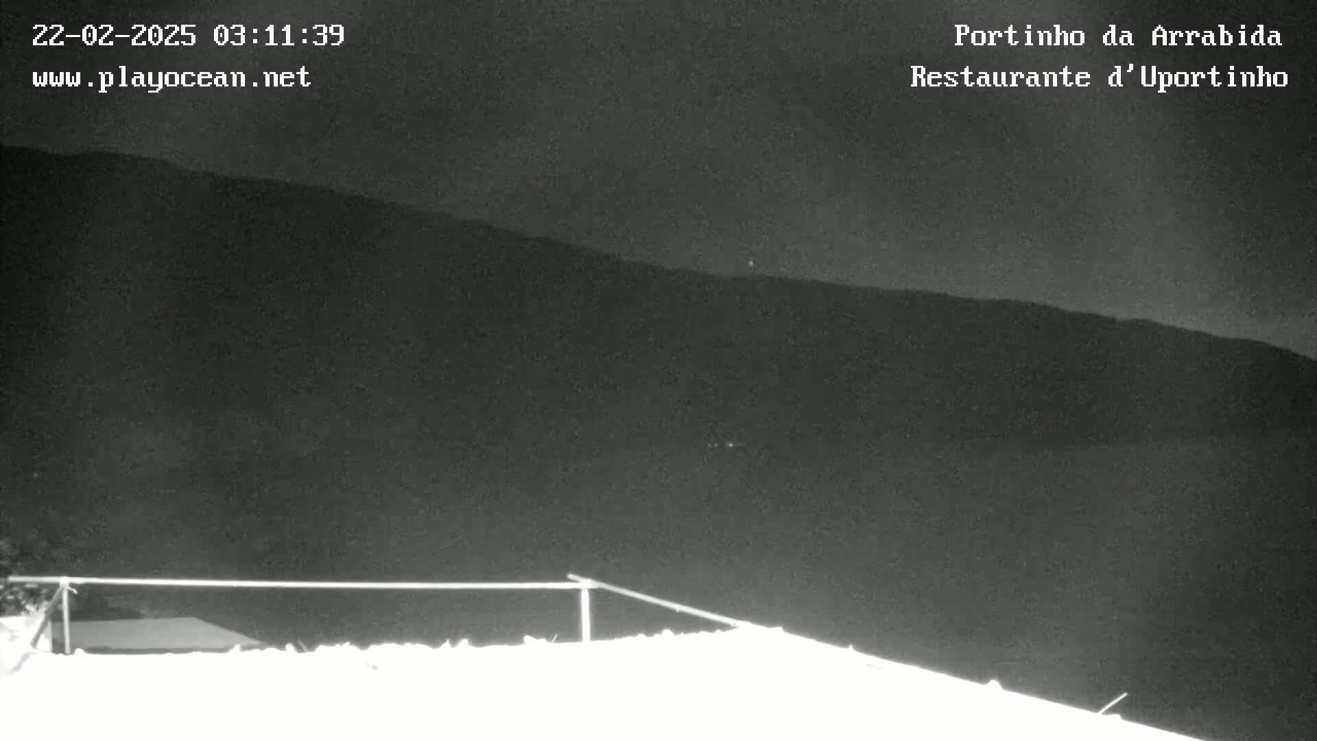 A nighttime, low-light image shows a snow-covered surface in the foreground with dark hills in the background under a clear, dark sky.