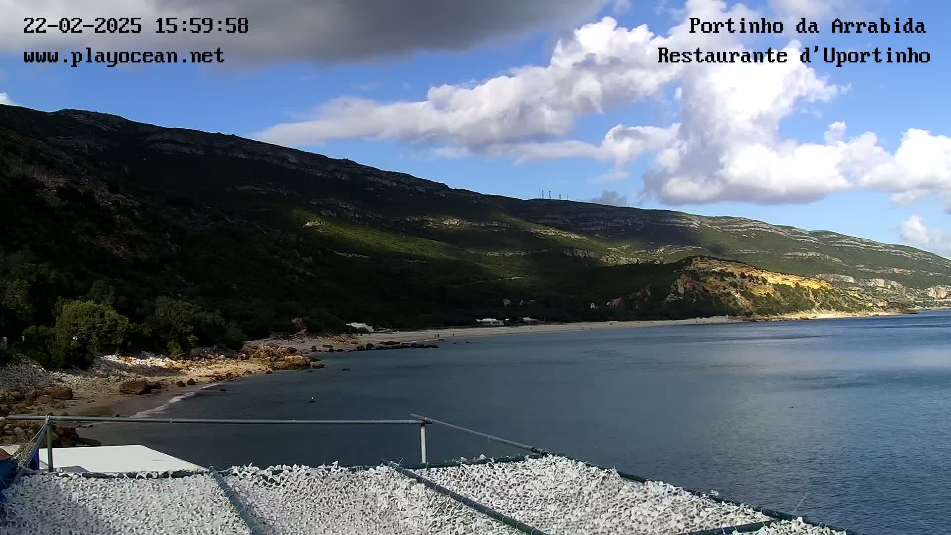 A partly cloudy day shows a view of a calm bay with a sandy beach and green hills in the background.