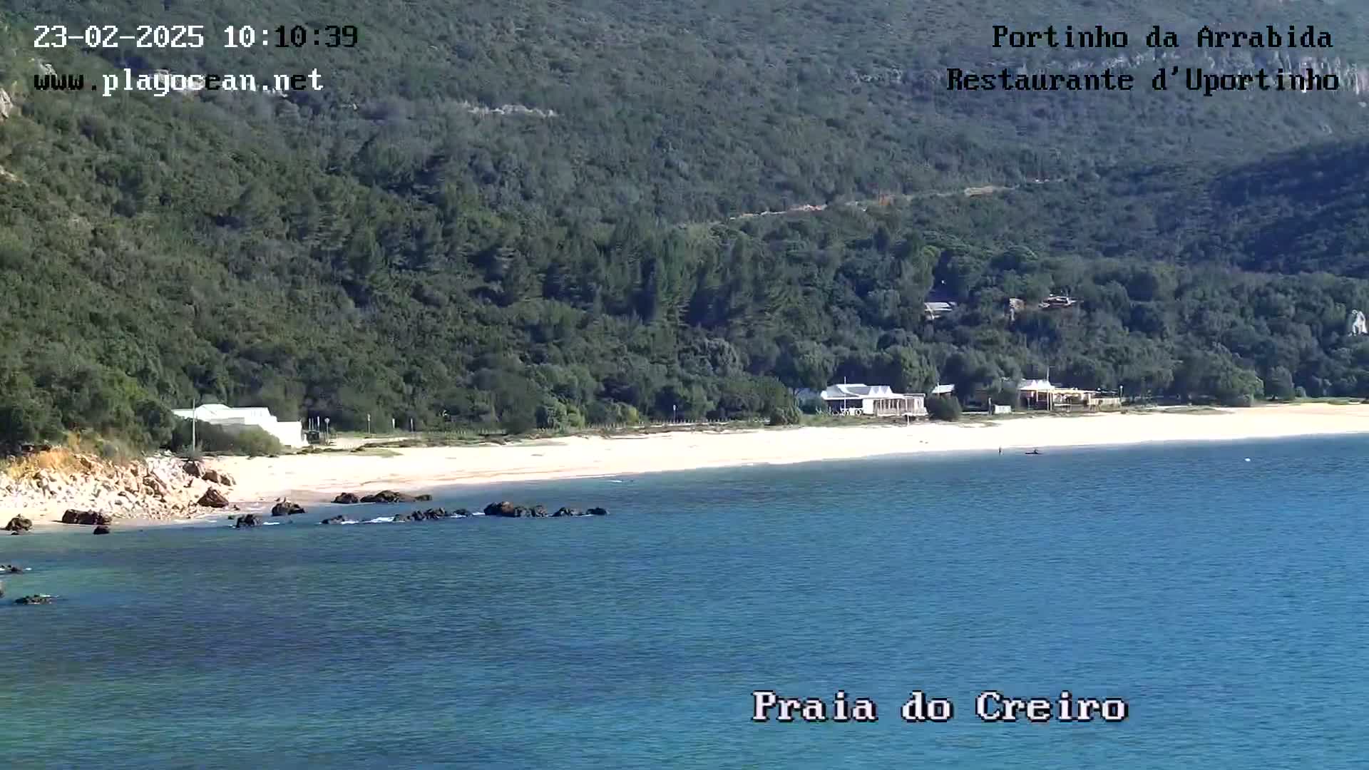 A sandy beach with calm blue water and several buildings is nestled against a lush green hillside on a sunny day.