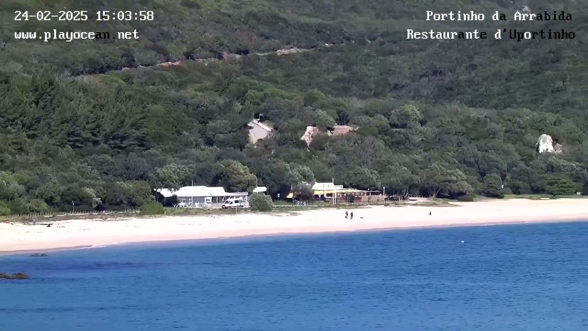 A sandy beach with a few people and buildings sits below a lush green hillside next to calm blue water.