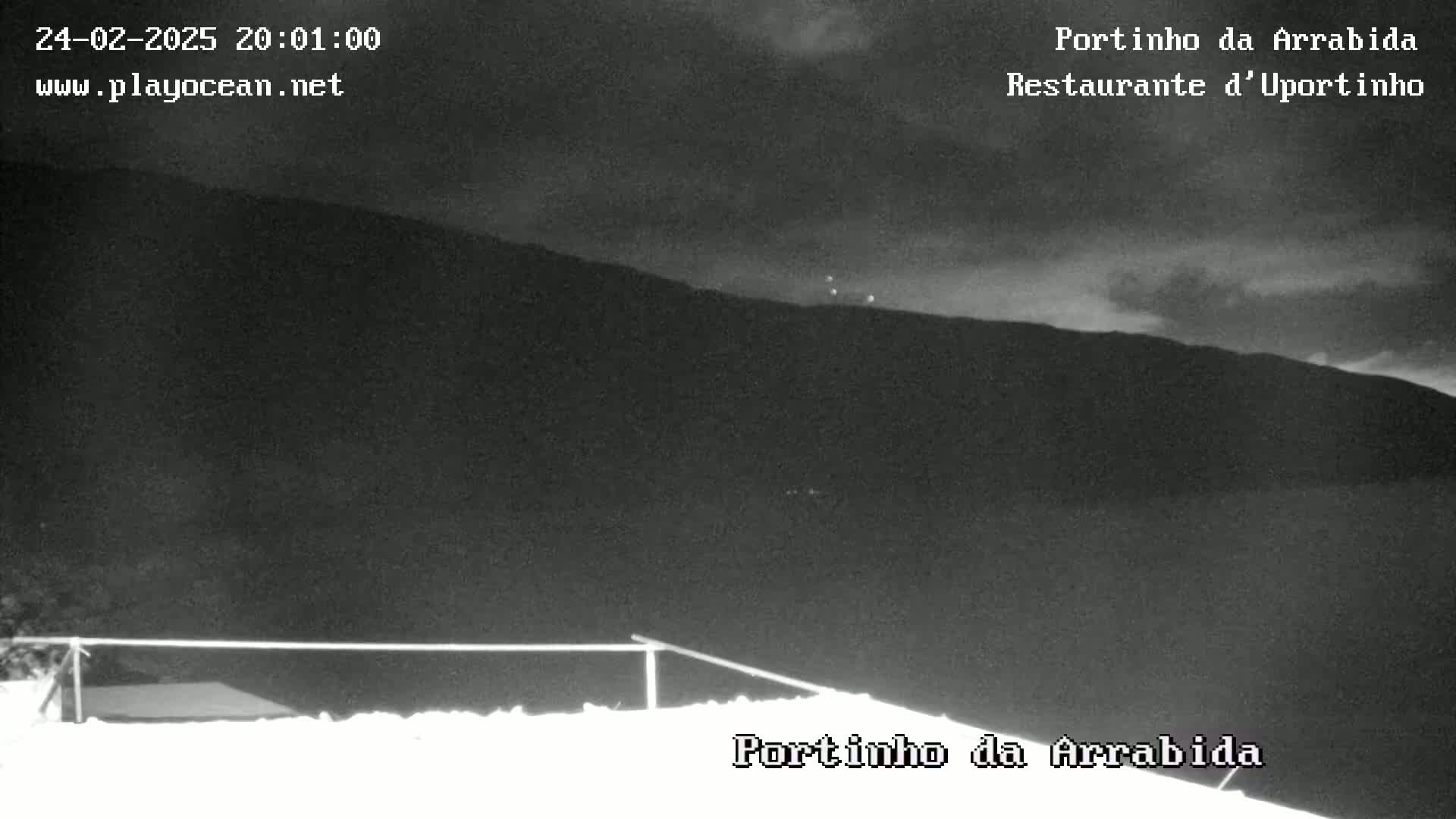 A nighttime, low-light view shows a dark hill in the distance, a low fence in the foreground, and a few faint lights on the hill.