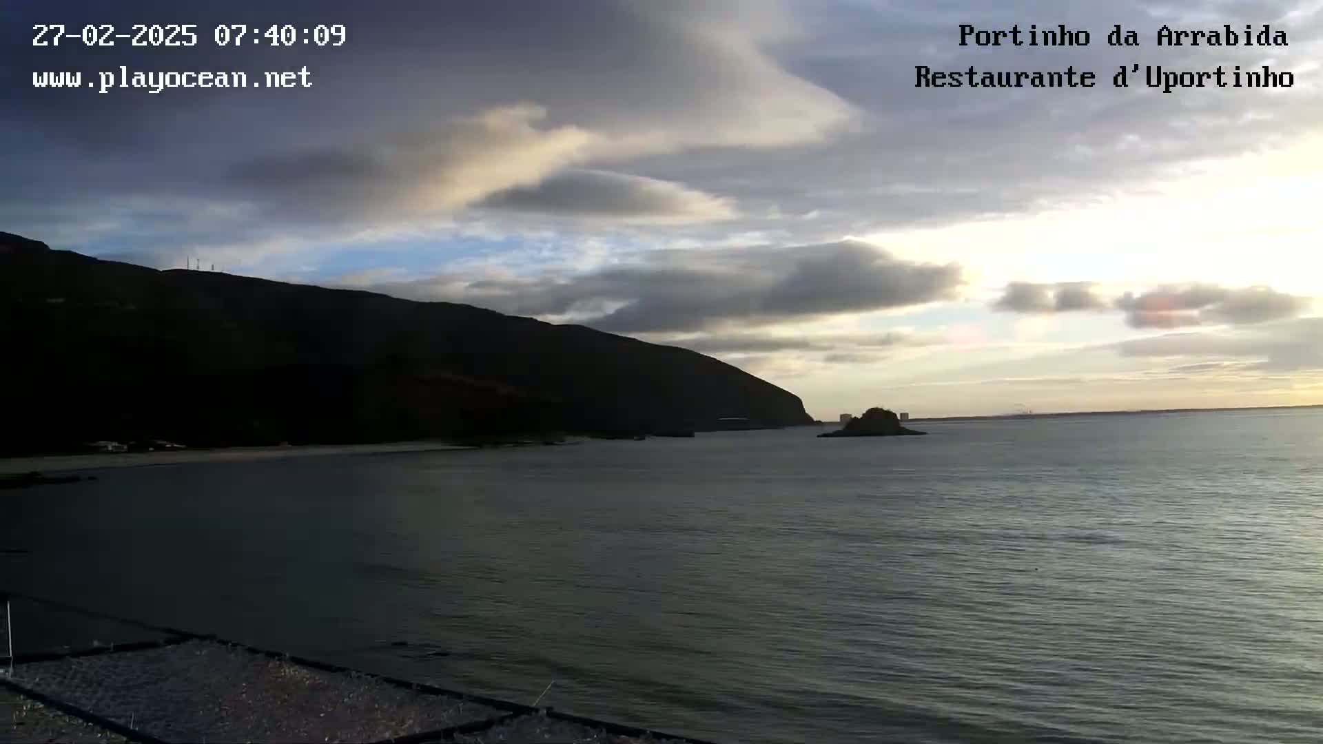 A partly cloudy sunrise illuminates a calm bay with a dark mountain in the background.