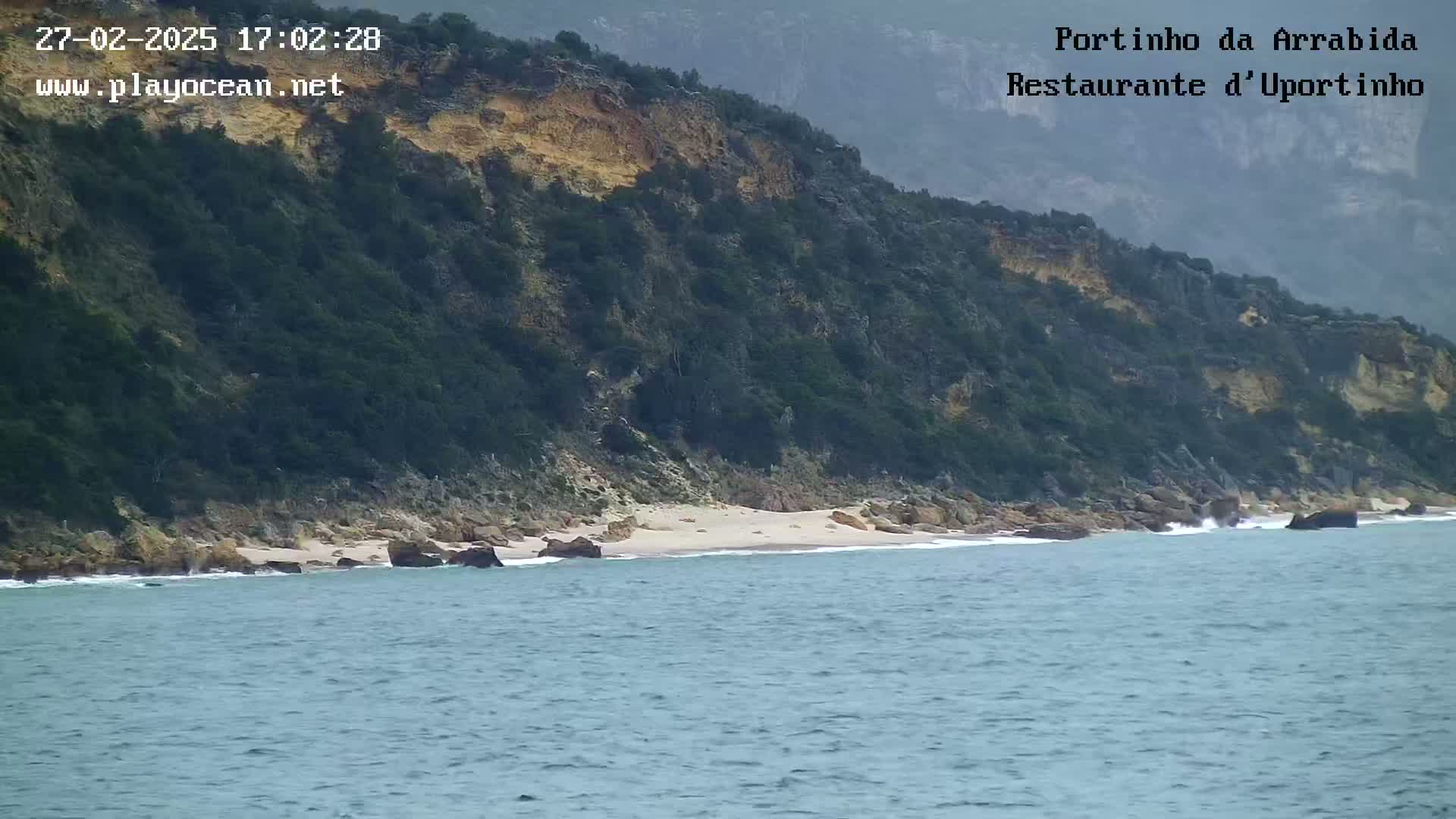 A rocky, vegetated cliff overlooks a small, sandy beach and calm ocean water.