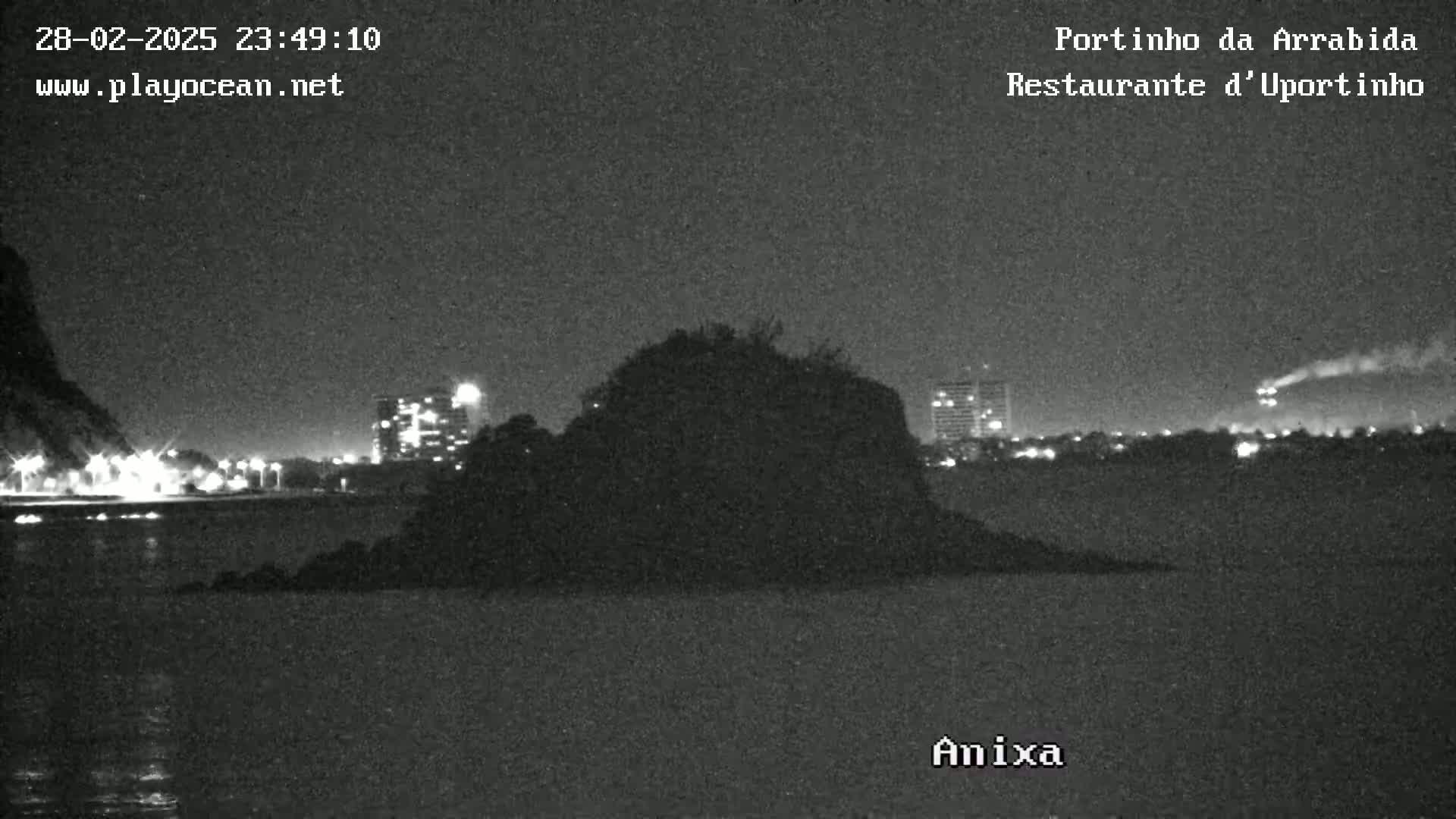 A dark, nighttime image shows a rocky island in calm water with city lights and buildings in the background under a clear sky.