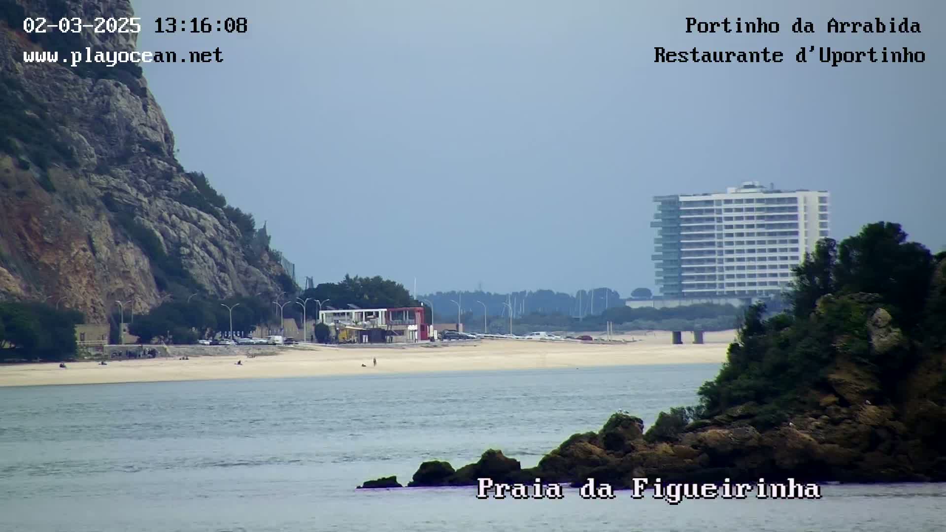 A sandy beach with a few people, a restaurant, and a tall building is visible across a body of calm water under a clear sky.