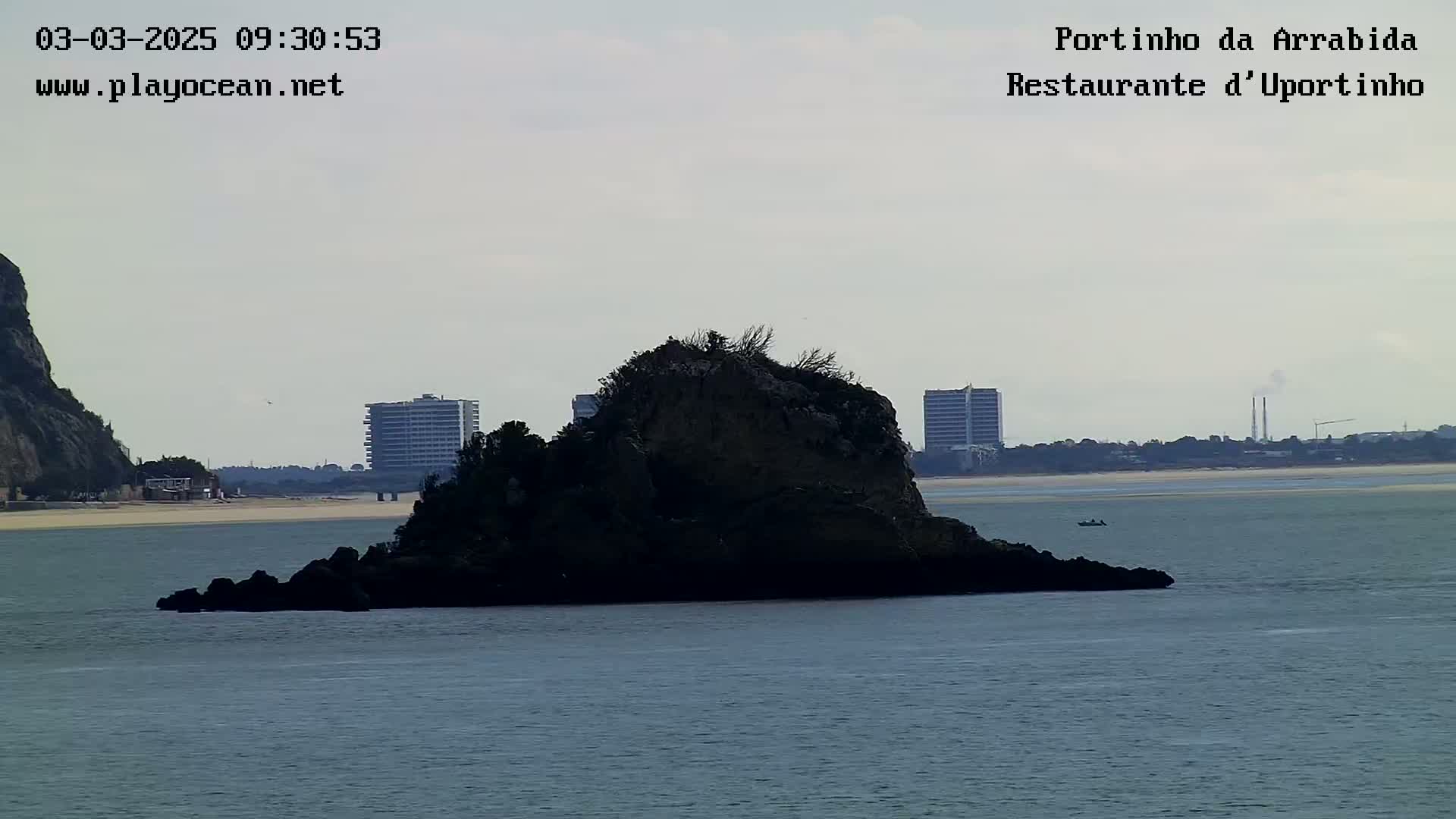 A rocky islet sits in calm, gray water in front of a coastline featuring buildings and a sandy beach under a cloudy sky.