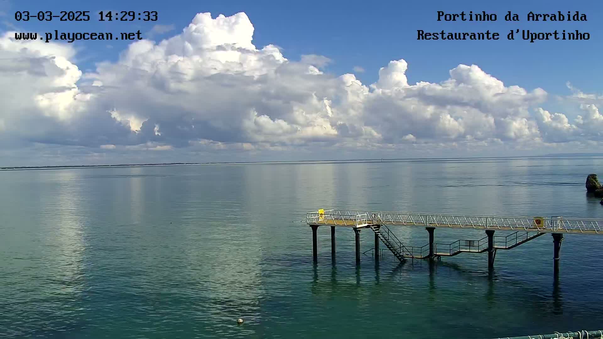 A calm, blue-green body of water stretches to a distant shoreline under a partly cloudy sky, featuring a small pier extending into the water.