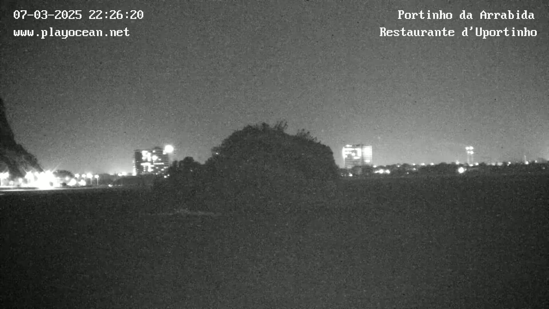 A nighttime, low-light view shows a dark, silhouetted landscape with city lights and buildings in the distance.
