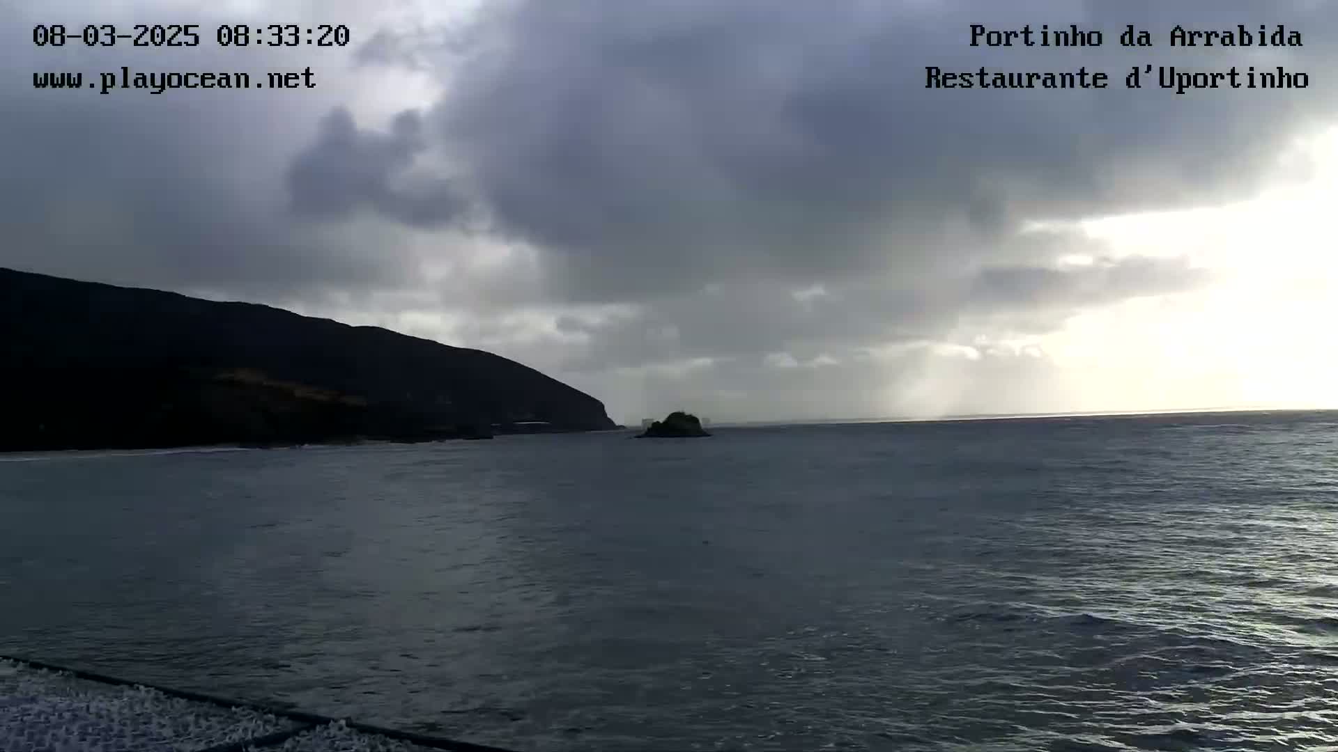 A dark, cloudy day shows a calm body of water with a small island and a dark mountain in the distance.