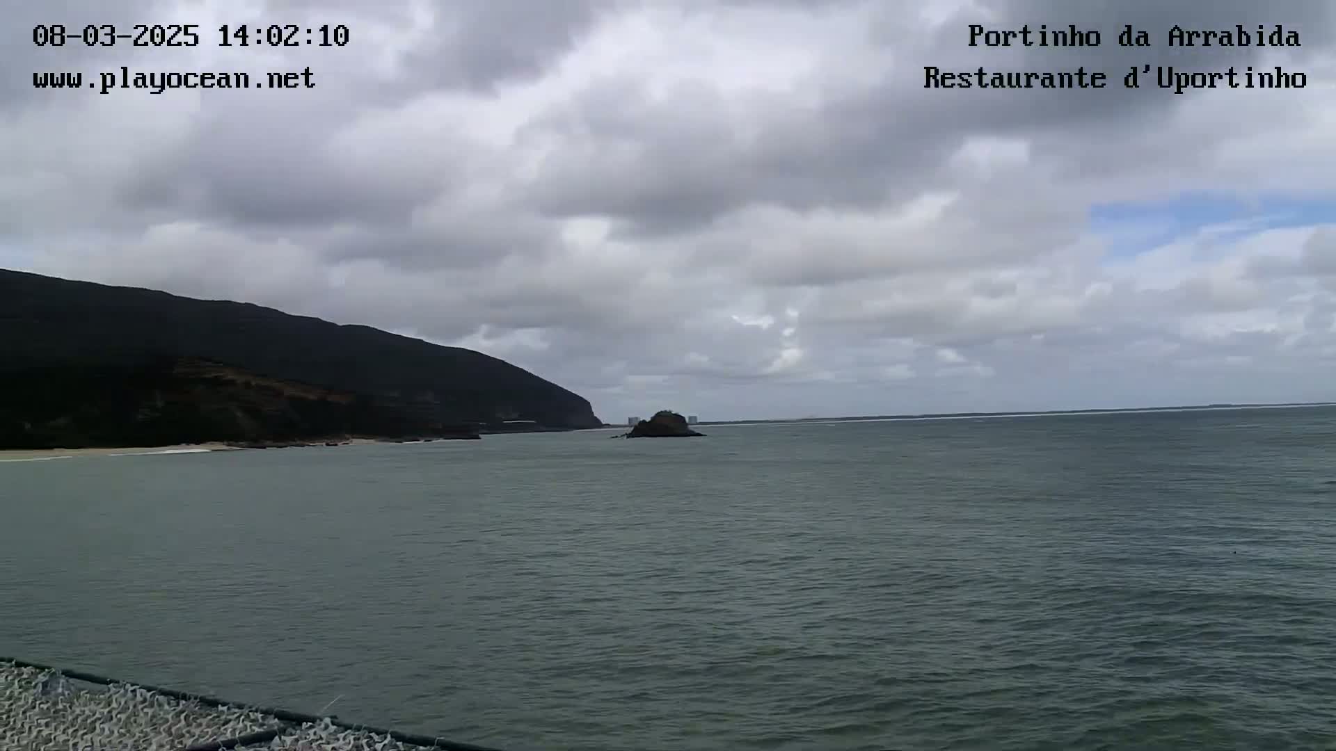 A dark, rocky coastline with a small island in the ocean under a mostly cloudy sky.