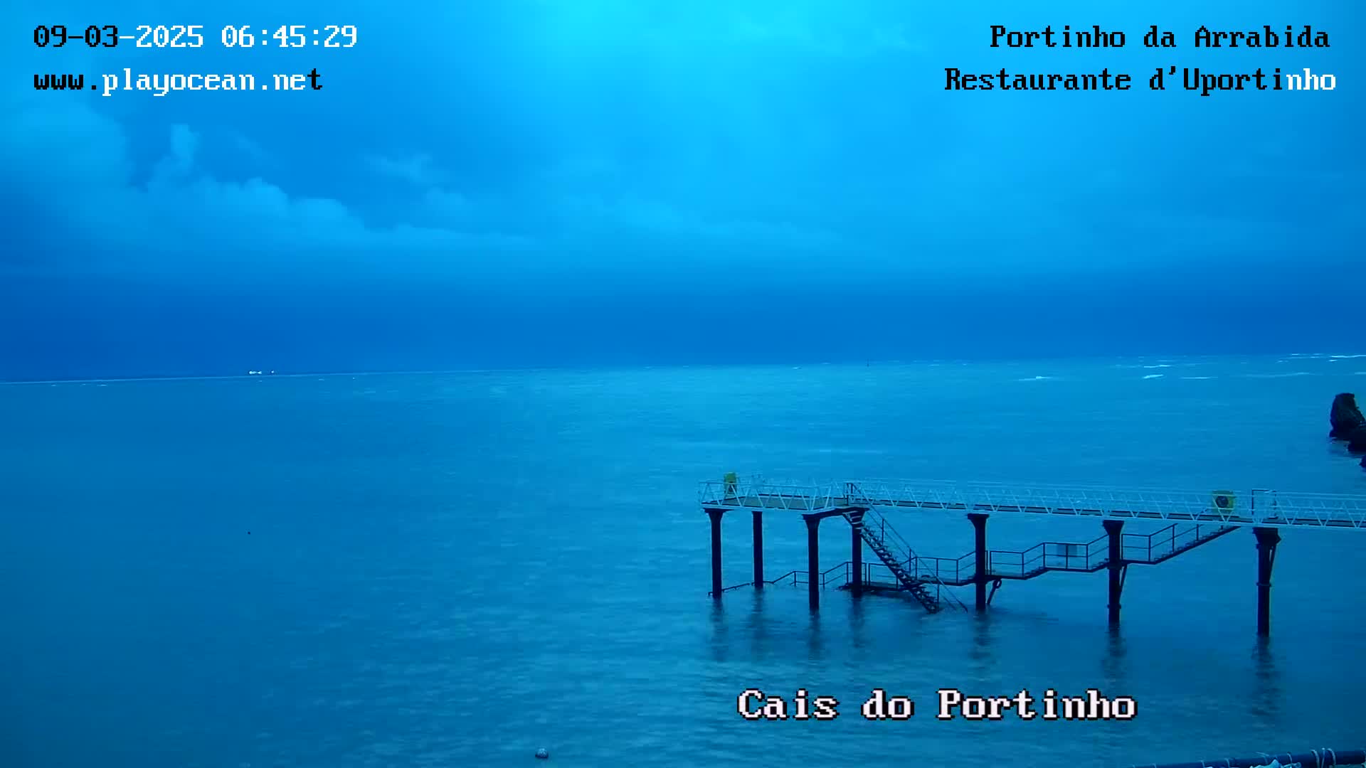 A pier extends into a calm, blue ocean under a mostly cloudy, dark blue sky.
