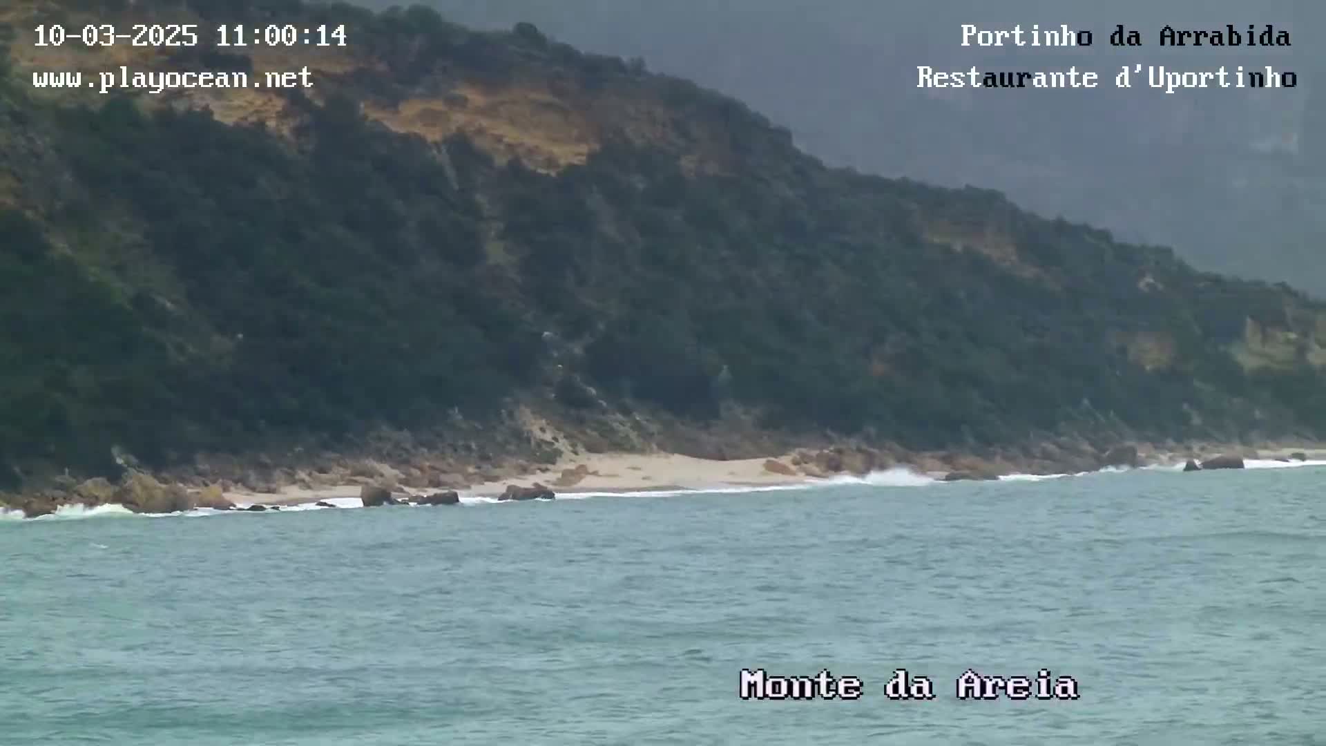A rocky, vegetated hillside meets a small, sandy beach and a moderately wavy ocean under an overcast sky.