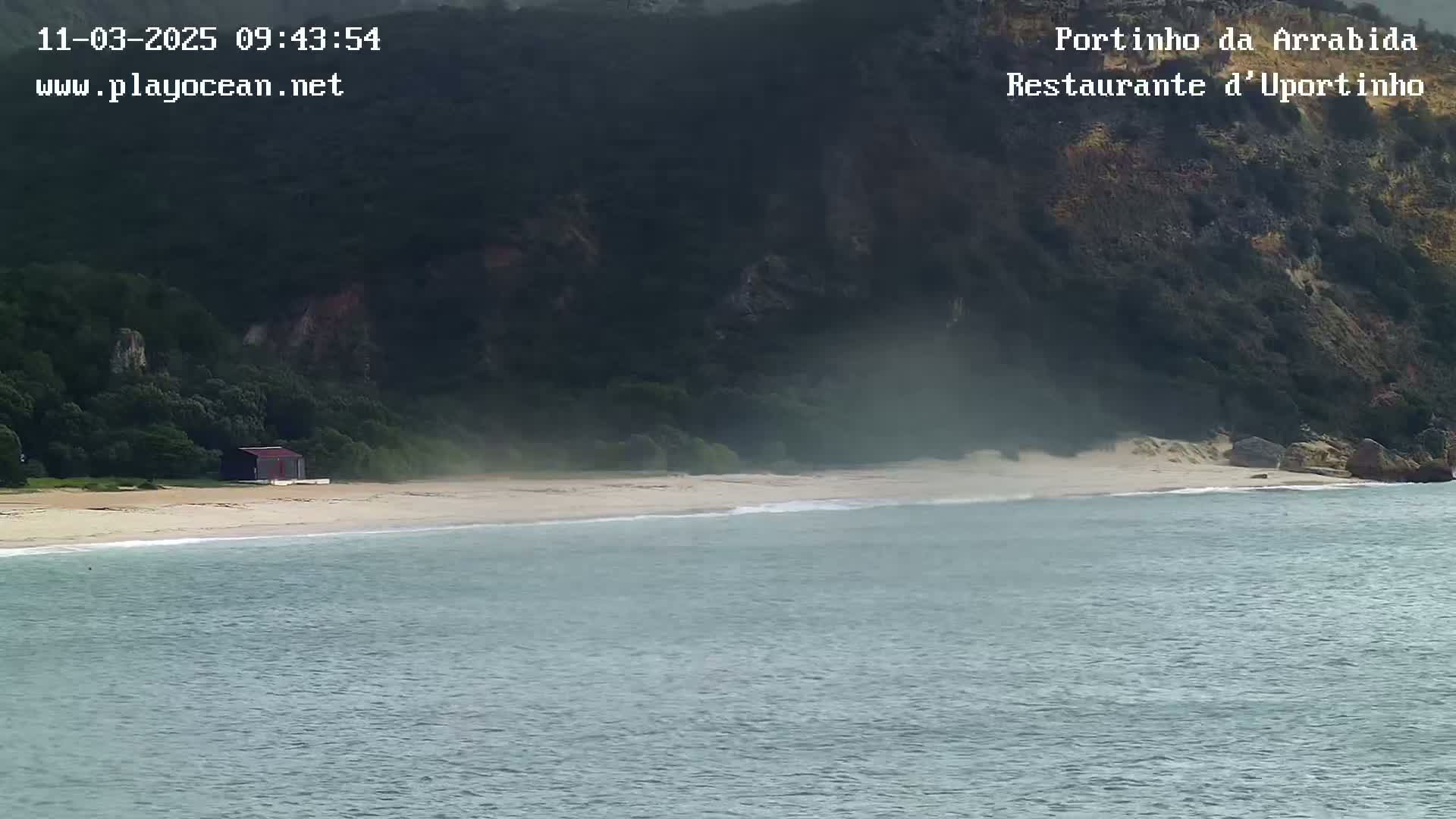 A sandy beach with a small building and calm ocean meets a dark green, steep cliff under an overcast sky.