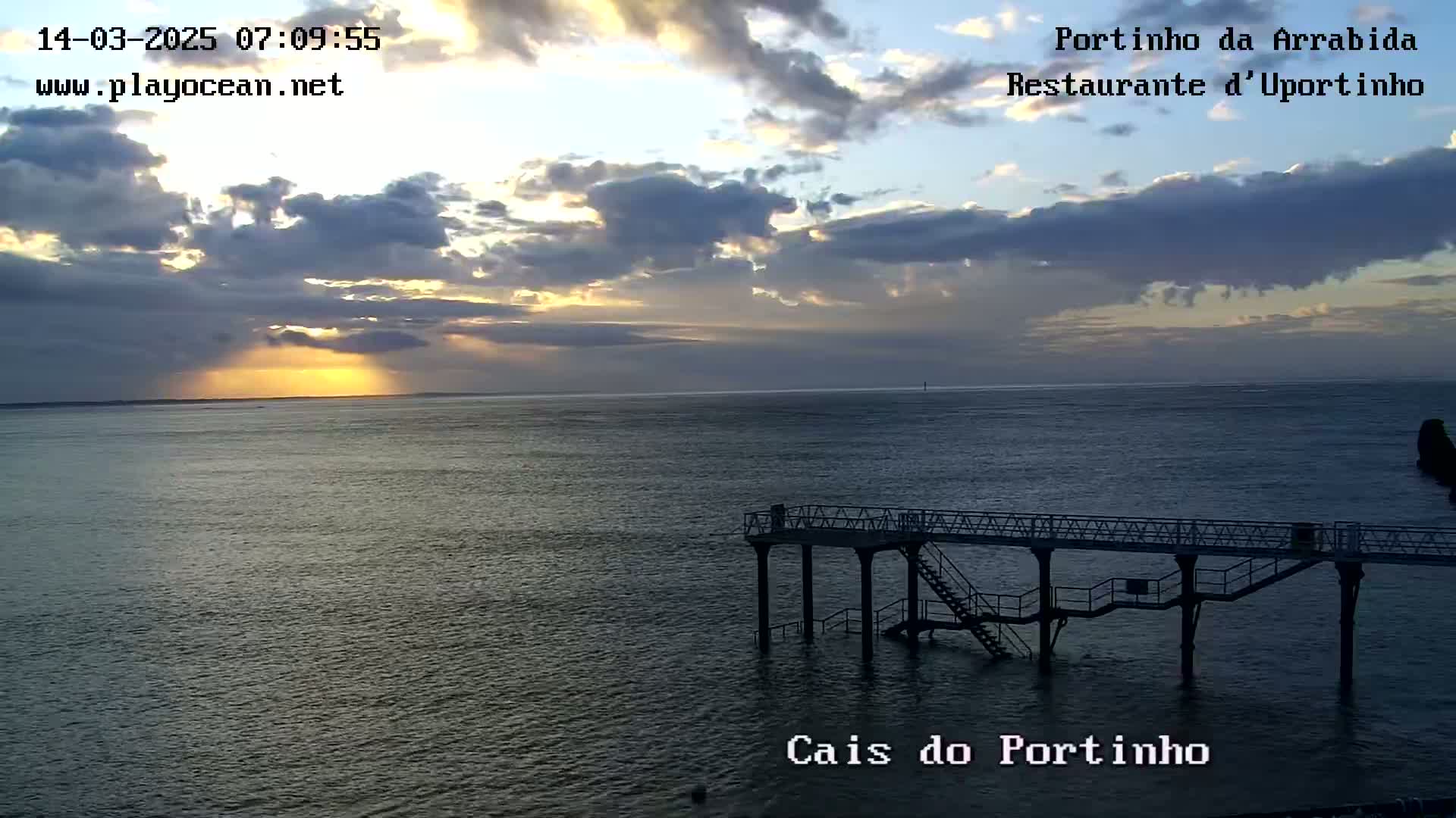 A partly cloudy sunrise illuminates a calm body of water where a pier extends into the sea.