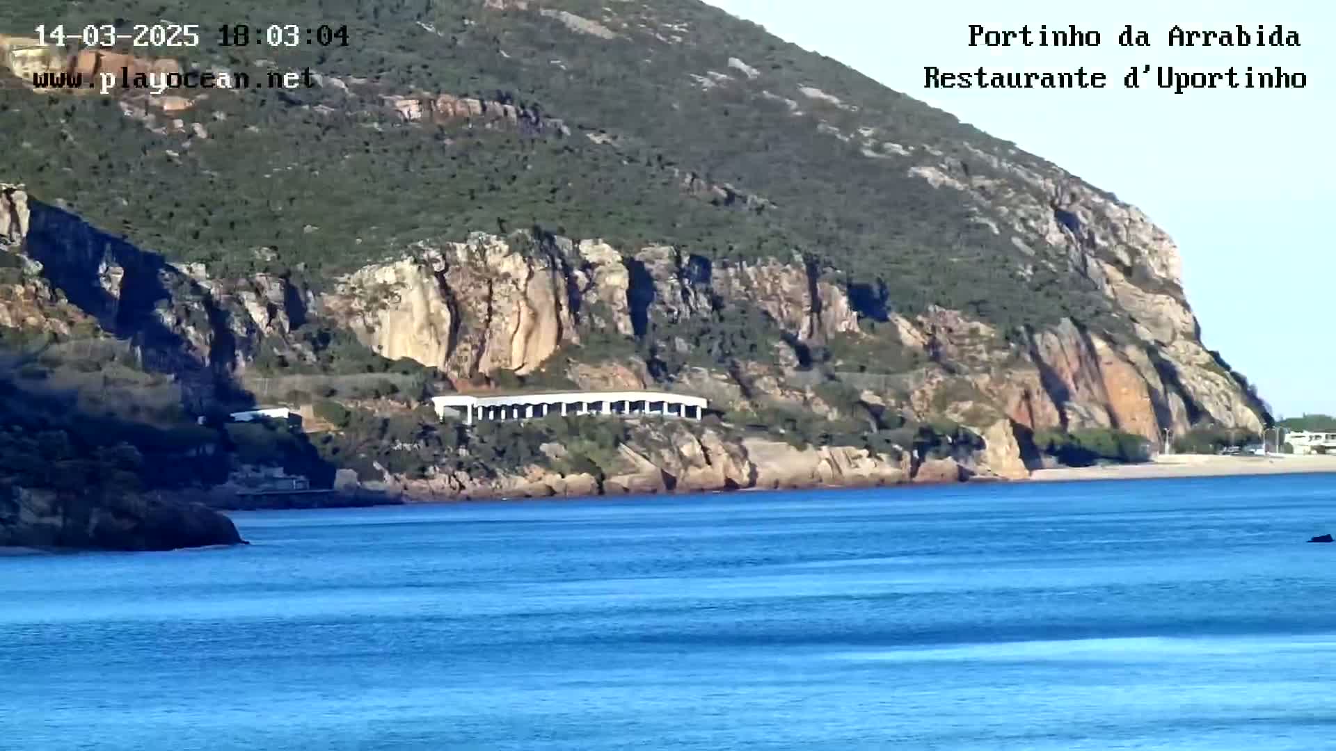 A long, low white structure is situated at the base of a rocky, green hill overlooking a calm blue ocean.