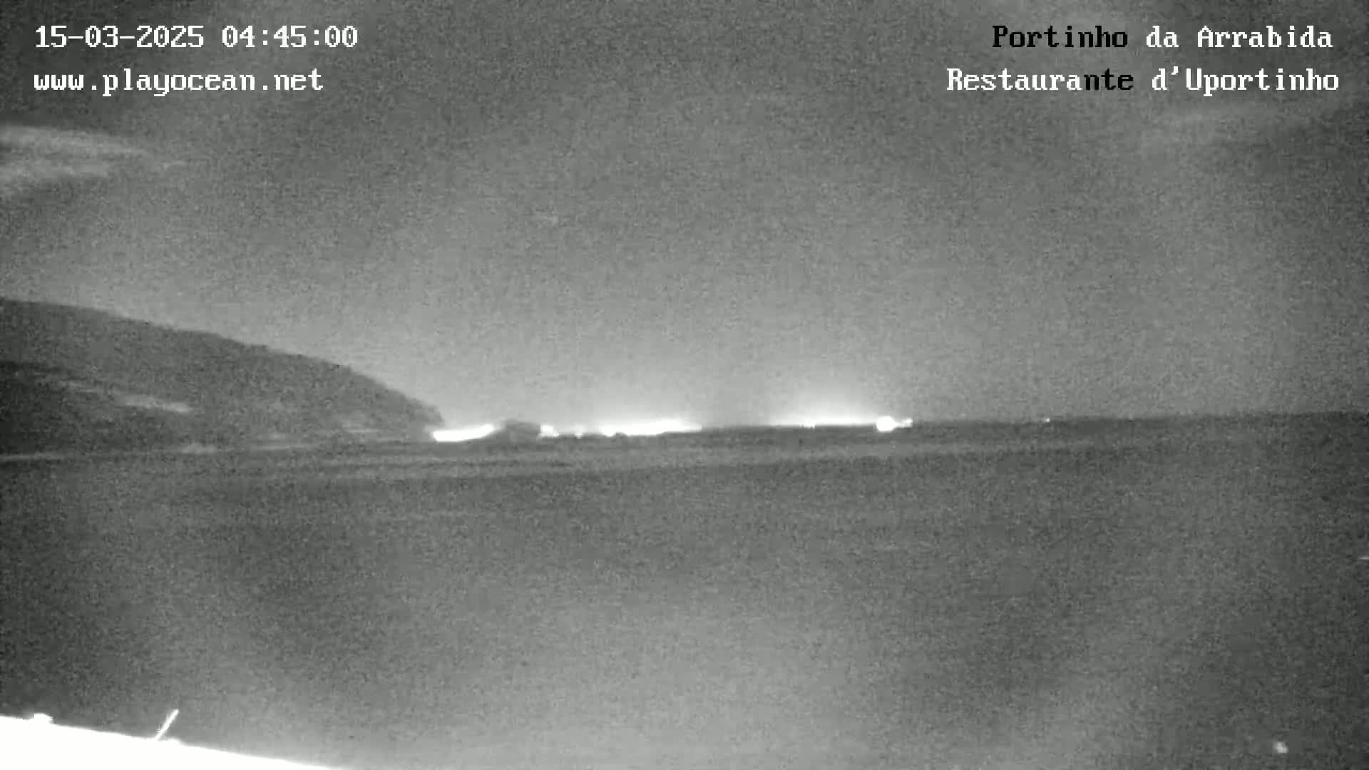 A nighttime, low-light image shows a dark body of water with a distant shoreline featuring faint lights, and a dark hill or mountain in the background under a mostly clear sky.