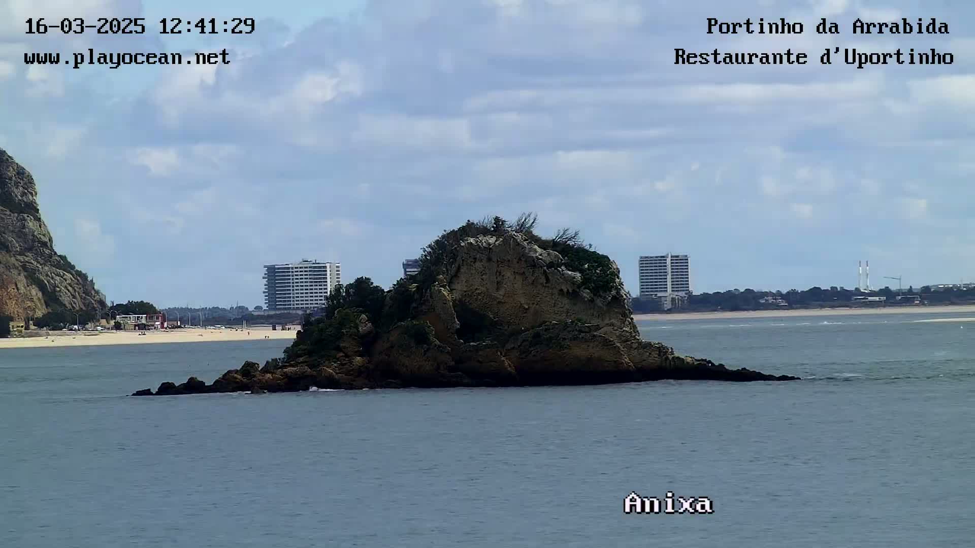 A rocky islet sits in calm, gray-blue water under a partly cloudy sky, with a sandy beach and distant buildings visible in the background.