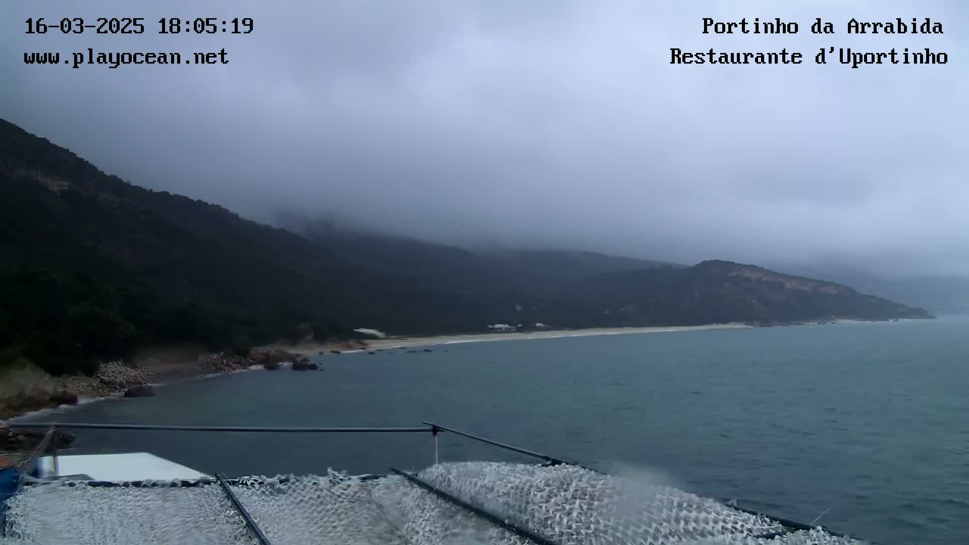 A view from a boat shows a hazy, overcast day over a calm bay with a sandy beach and dark green hills.