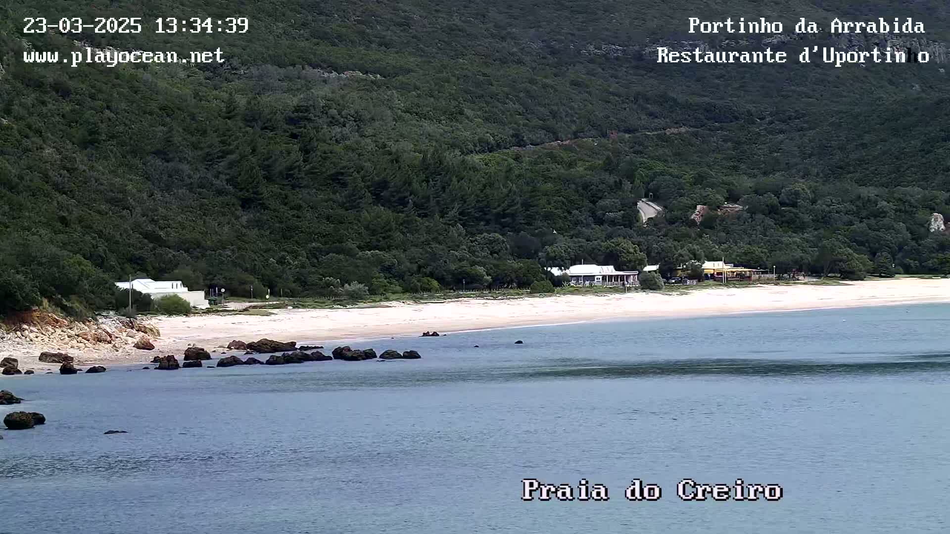 A sandy beach with calm water is nestled below a lush green hillside on a partly cloudy day.
