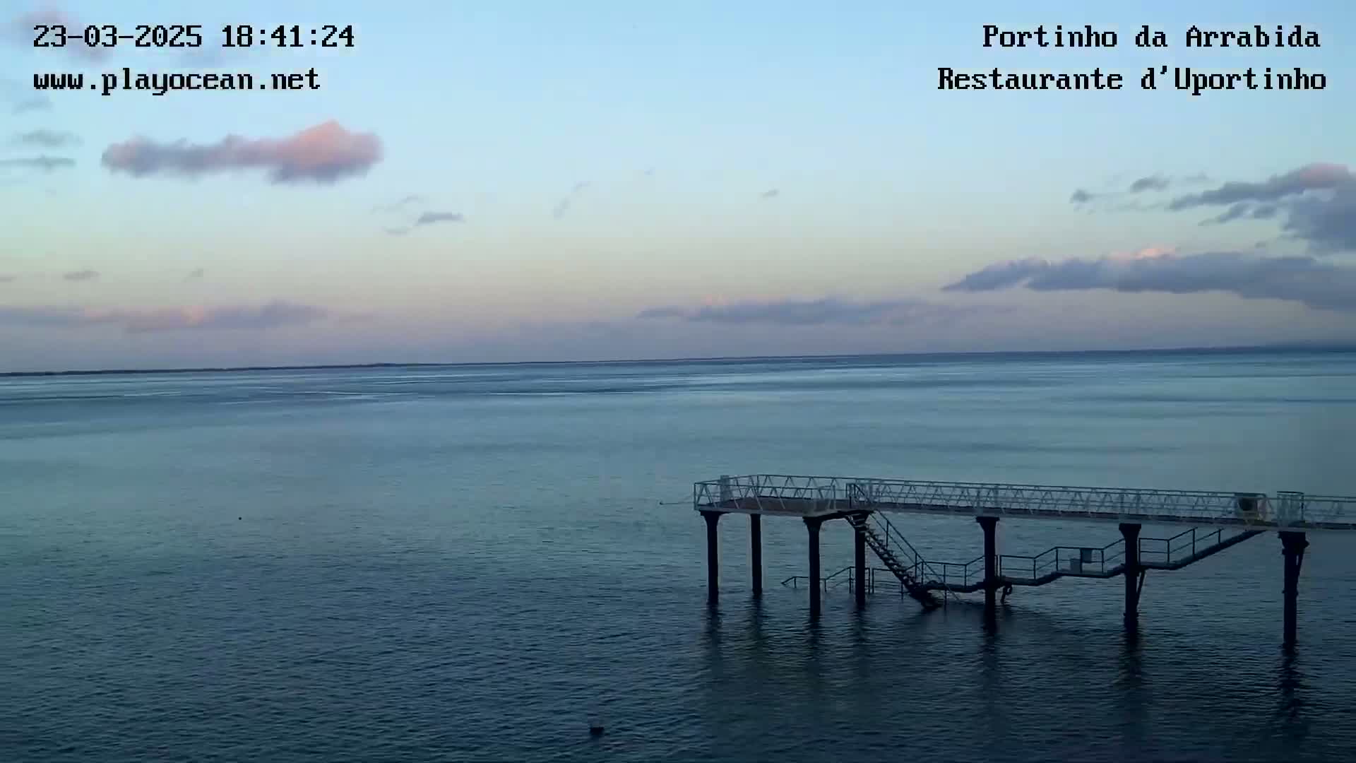 A calm ocean under a partly cloudy, pale pink and blue sunset sky features a small pier extending into the water.