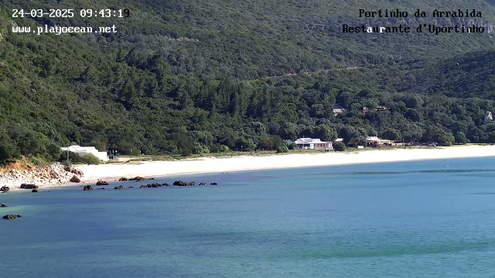 A sandy beach with calm, light-blue water is backed by a green, hilly landscape with a few buildings.