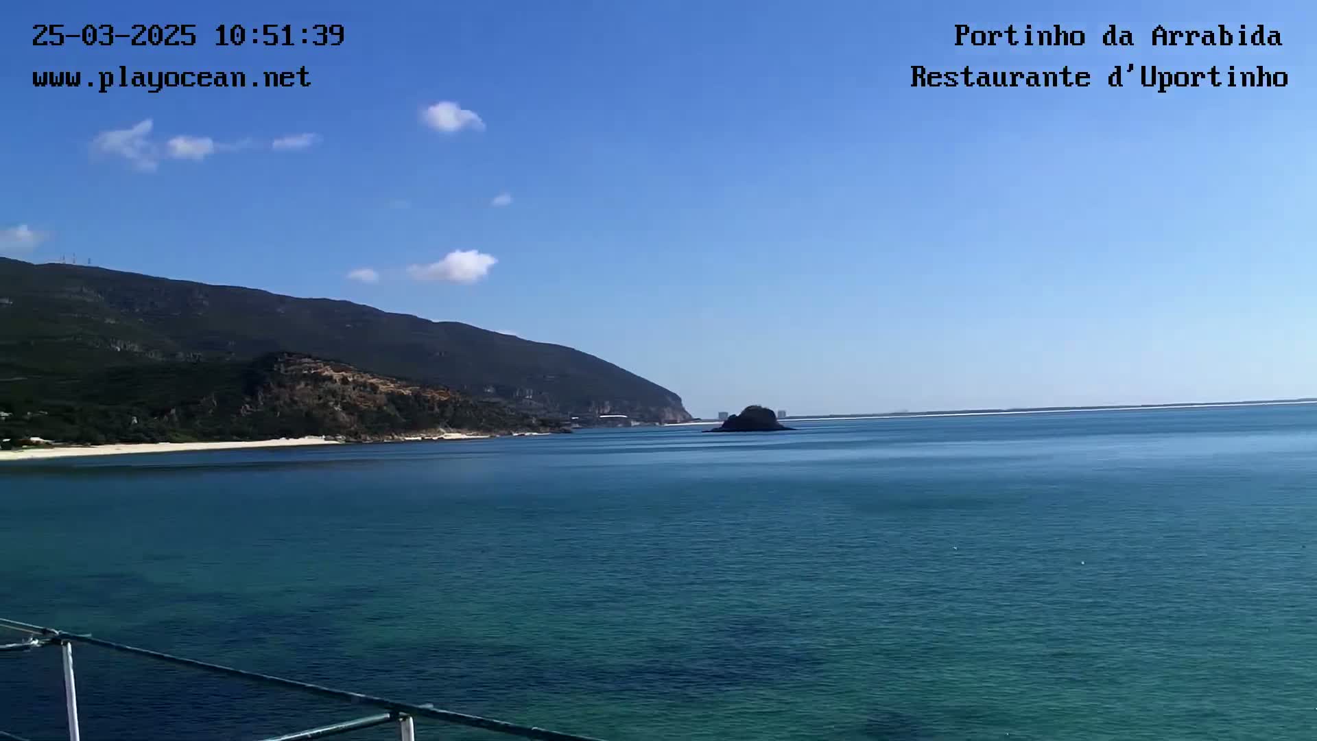 A tranquil coastal scene on a sunny day shows a calm, teal-colored ocean stretching toward a mountainous shoreline with a small island in the midground.