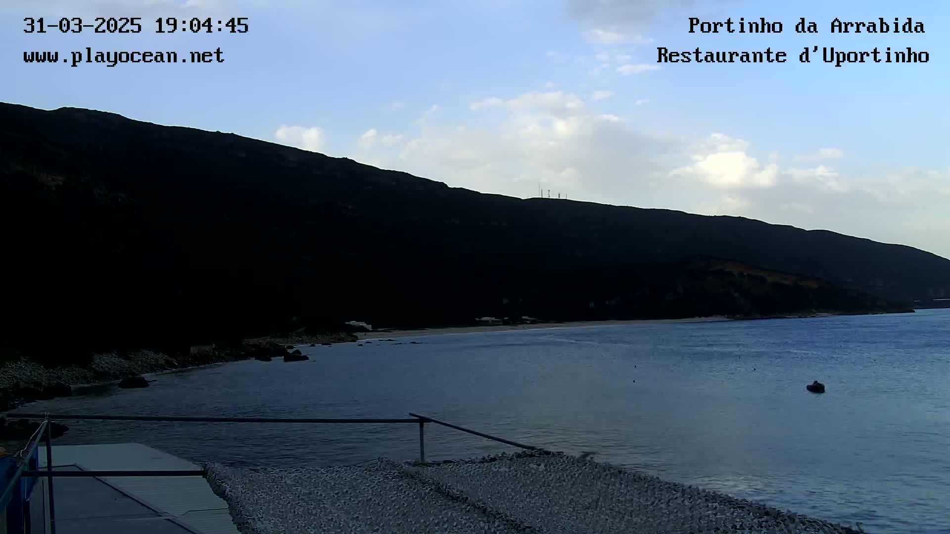 A calm, twilight ocean cove is nestled beneath a dark, silhouetted hill under a partly cloudy sky.