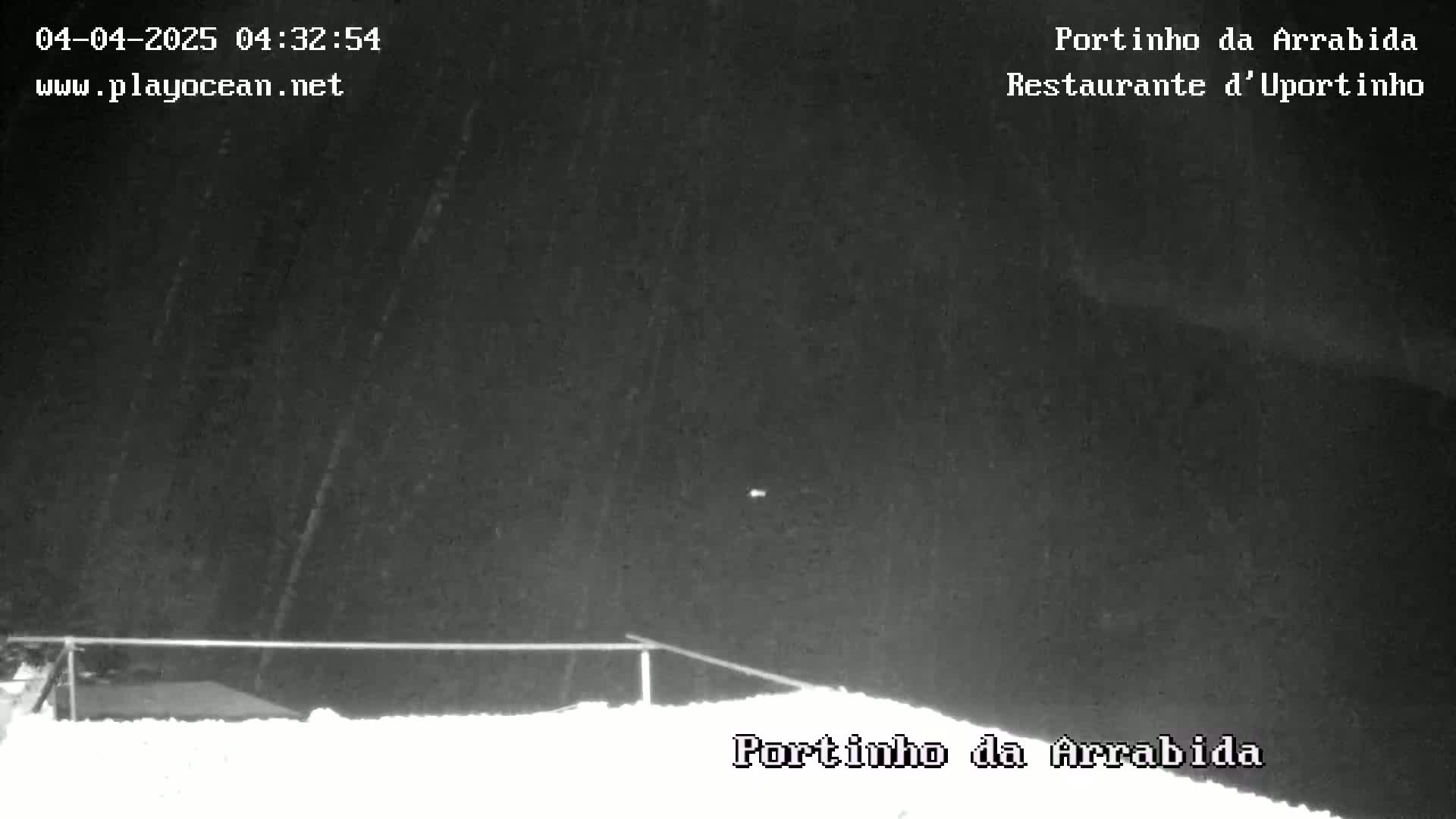 A nighttime, snowy scene shows a low-lying fence and a dark, overcast sky with light snowfall.