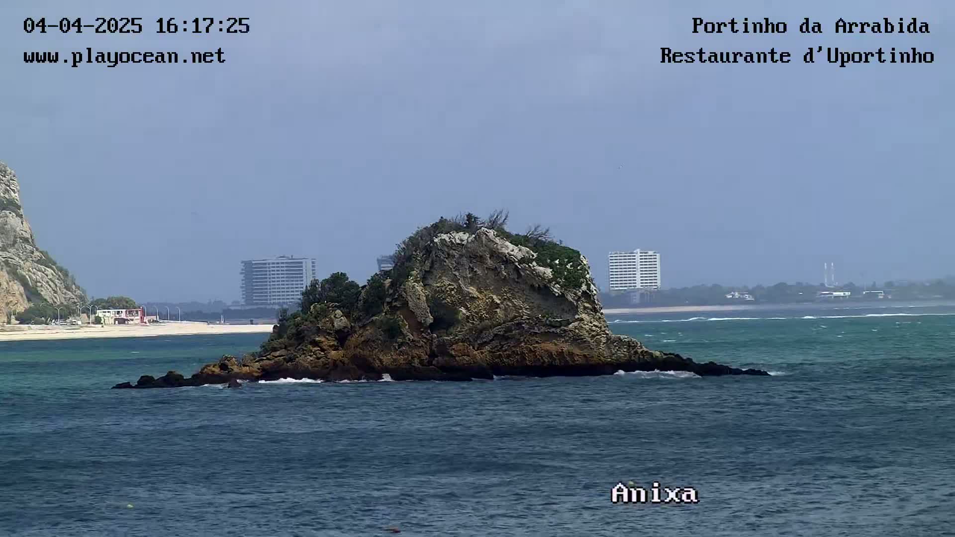 A rocky islet sits in a calm ocean near a sandy beach and distant buildings under a hazy sky.