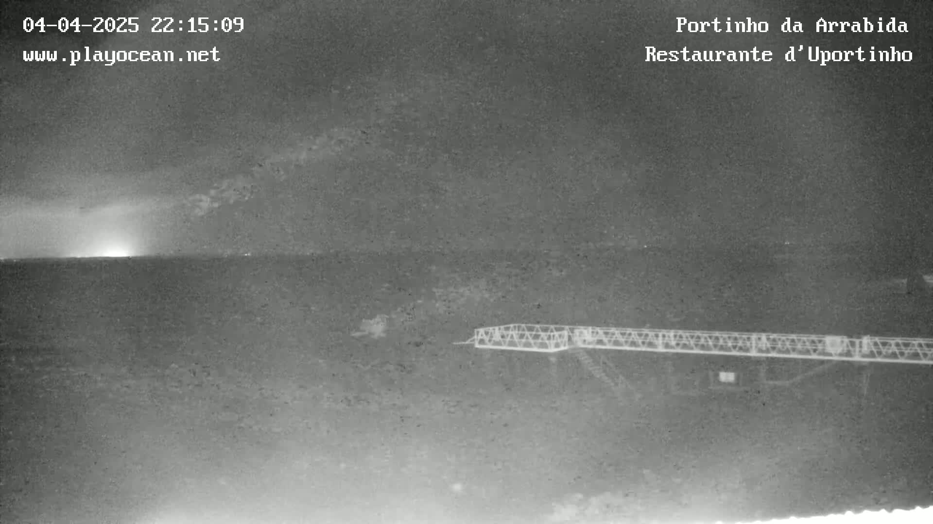 A nighttime, low-light image shows a metal structure extending over dark land or water under a mostly clear sky with a faint light horizon.