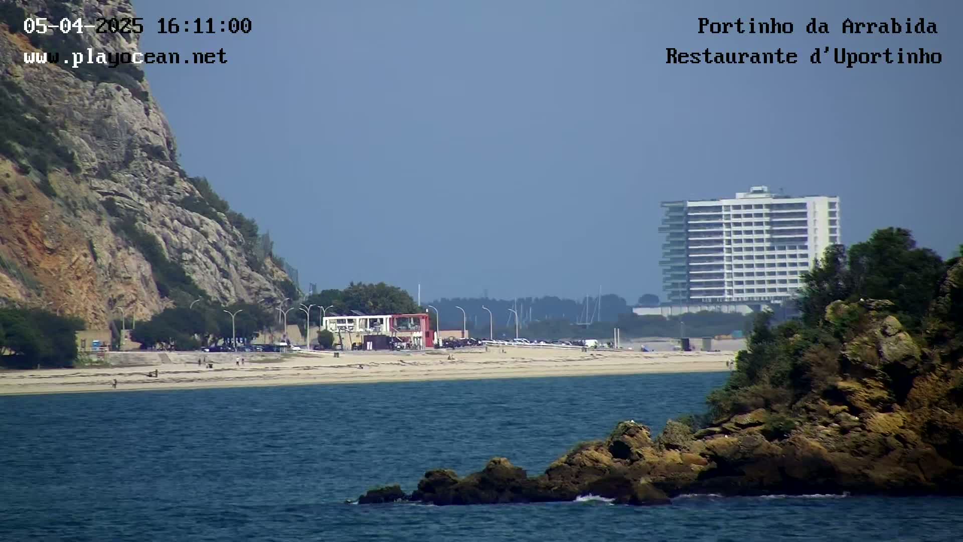 A sandy beach with a few people on it is situated between a rocky cliff and a large building, all under a clear sky.