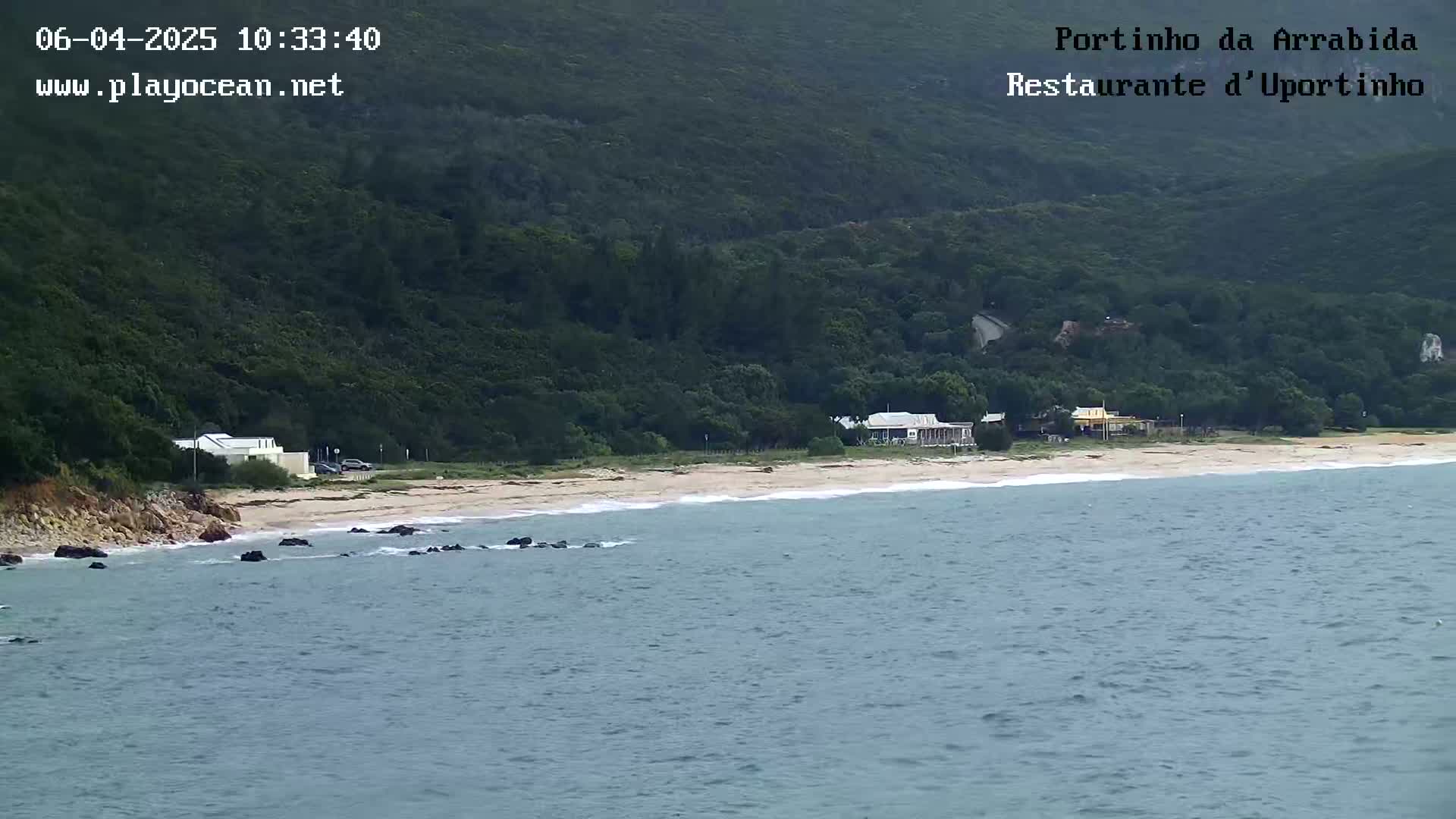 A sandy beach with several buildings sits below a lush, green hillside overlooking a calm, blue ocean.
