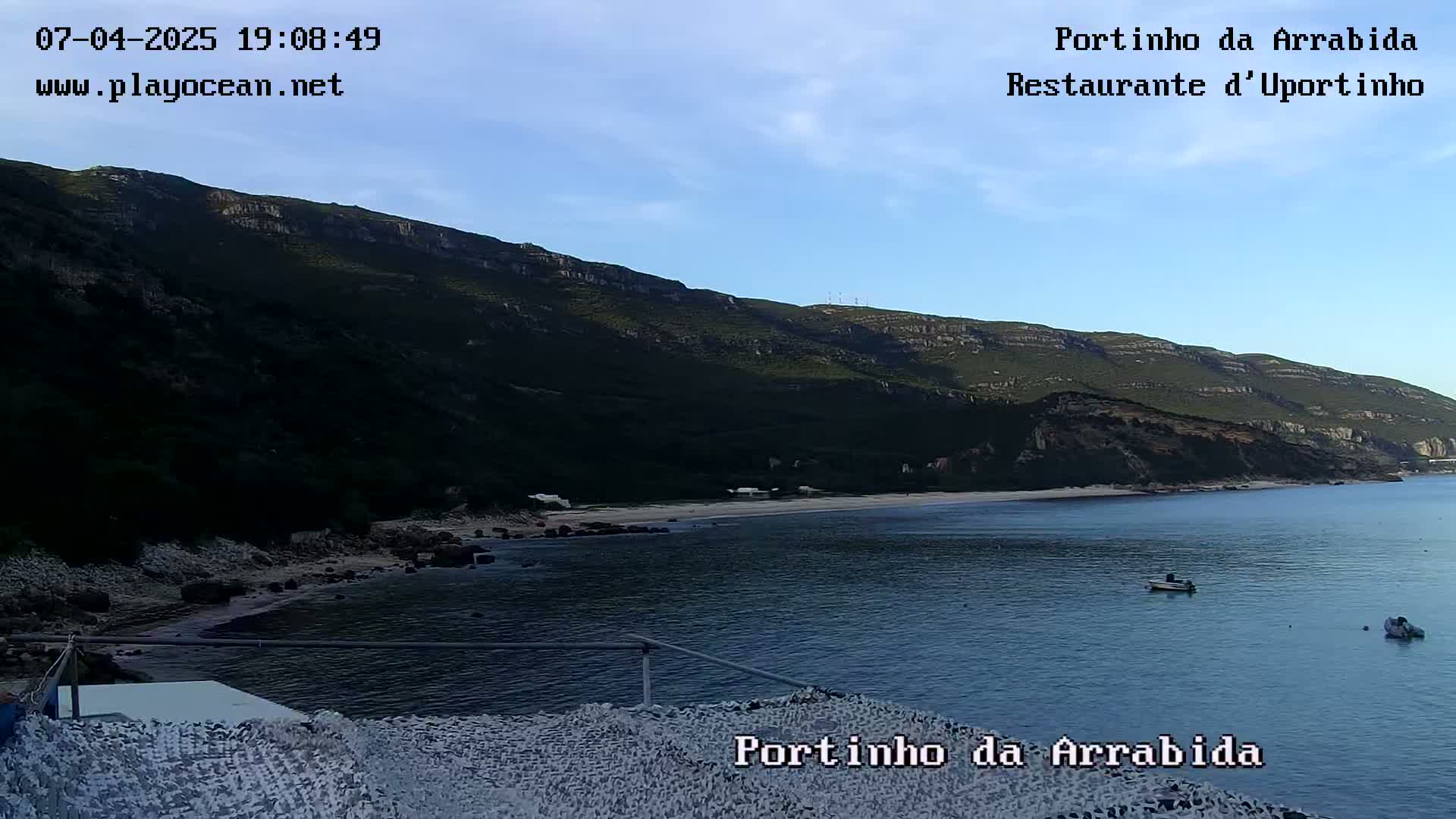 A tranquil cove with a small sandy beach nestled beneath a dark green mountain, under a clear blue sky.