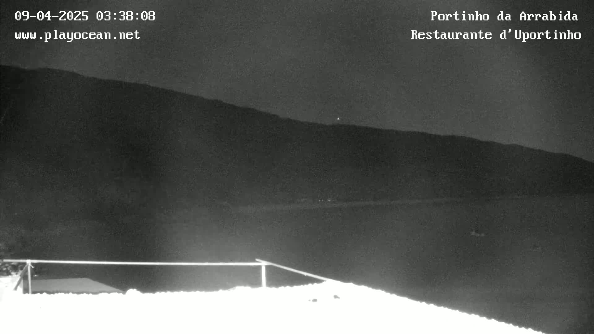 A nighttime, low-light view shows a dark mountain range in the distance, with a snow-covered surface in the foreground and a simple railing.