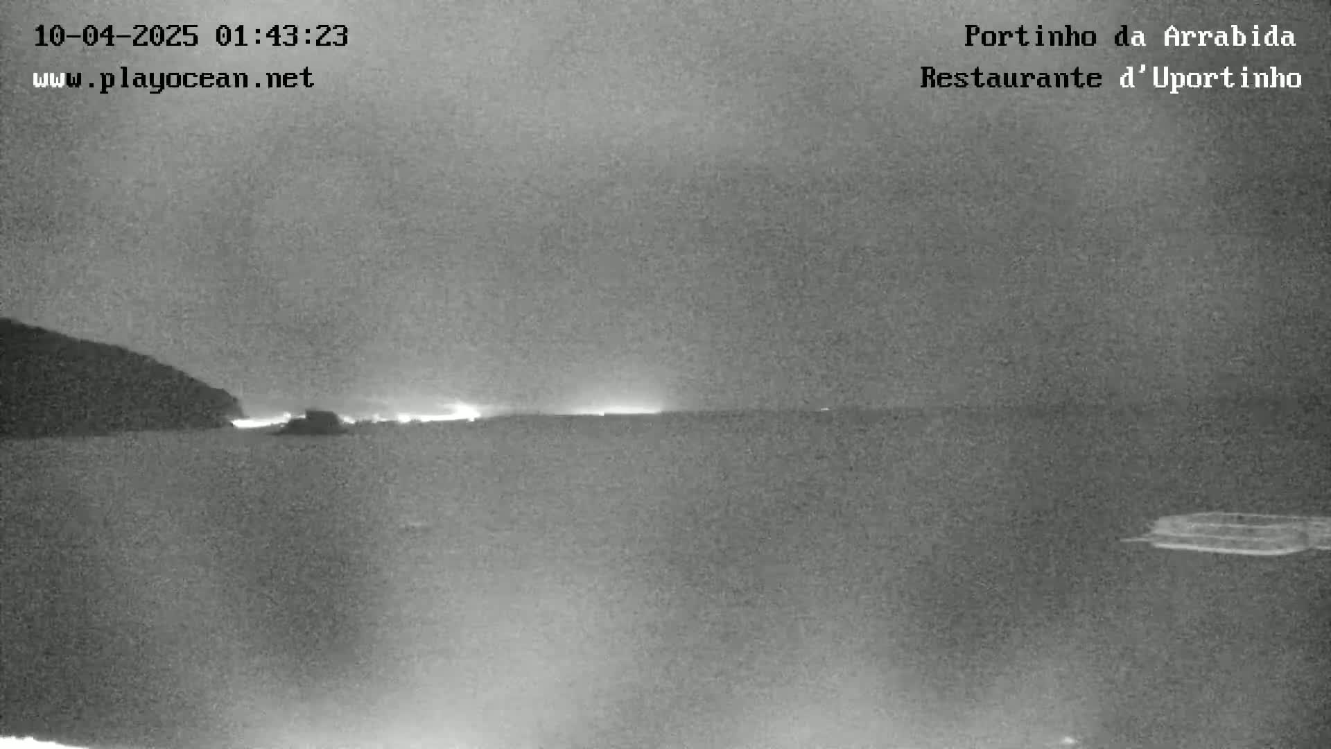 A nighttime, grayscale image shows a dark body of water with a faint horizon line and a dark landmass to the left, illuminated by some distant lights.