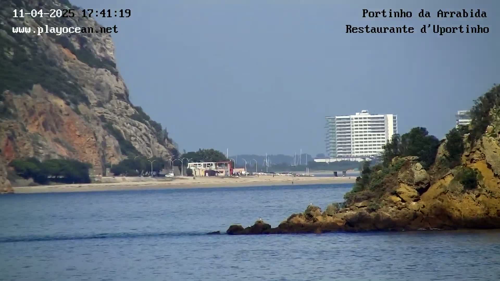 A sandy beach with a building and a rocky cliff is visible across a calm body of water, under a hazy sky.