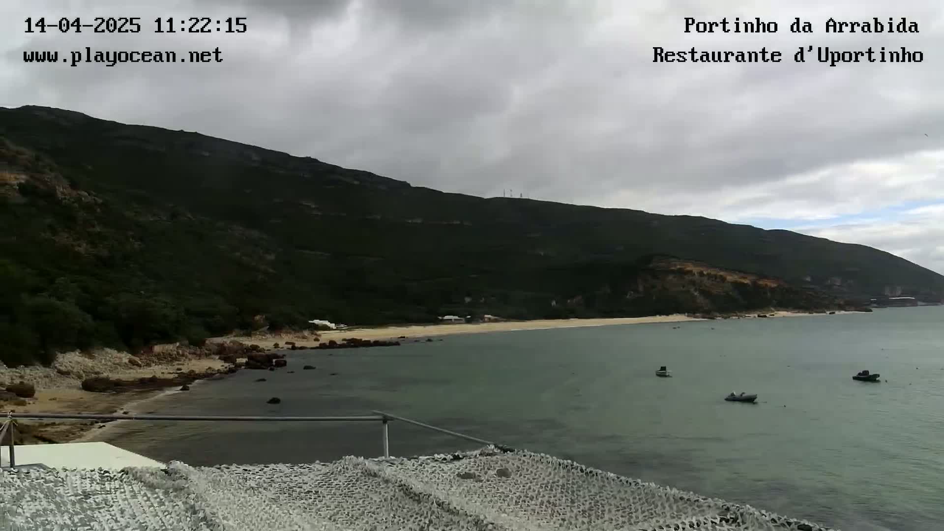 A view of a calm bay with a sandy beach and several small boats, overlooked by a dark green mountain under a cloudy sky.