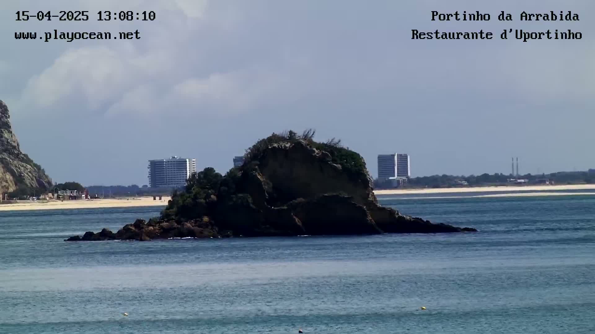 A rocky island sits in calm ocean water, with a sandy beach and distant buildings visible in the background under a mostly cloudy sky.