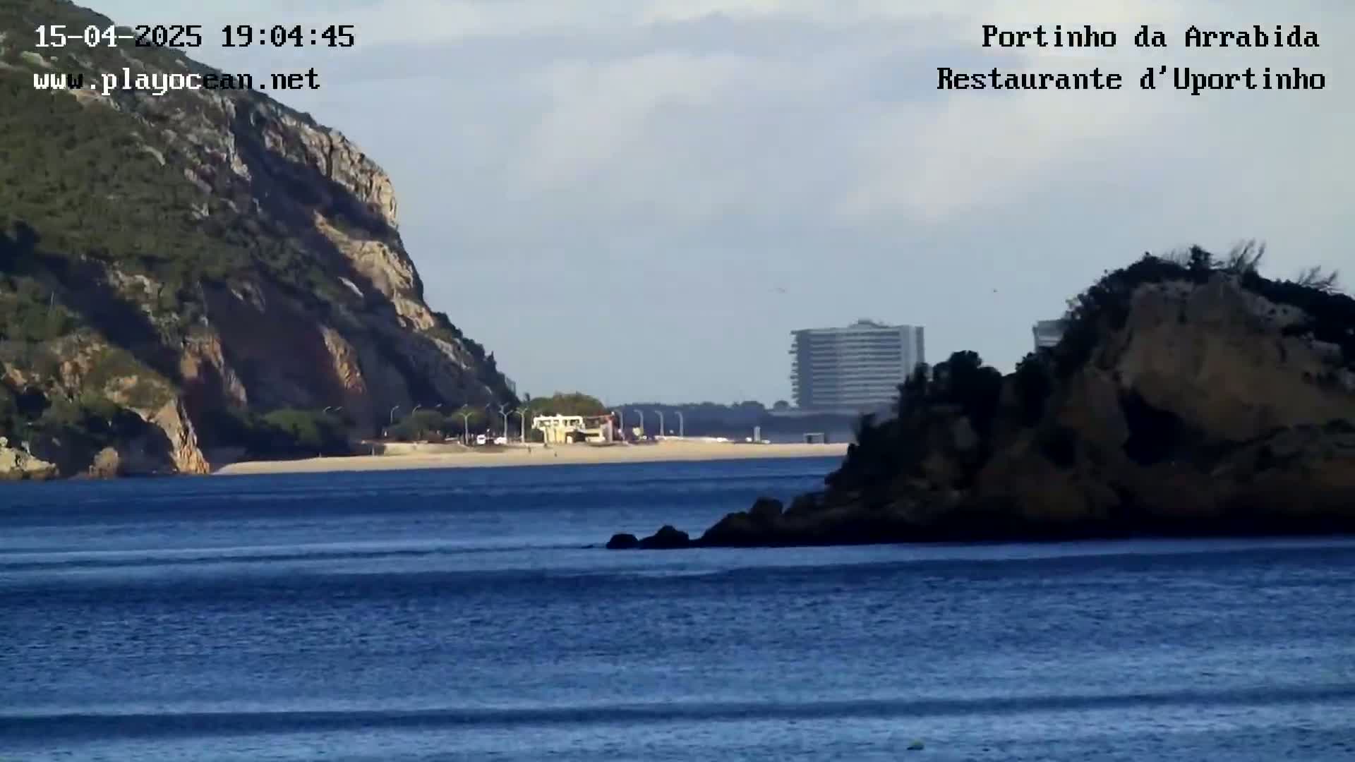 A calm, blue body of water separates a rocky, vegetated shore from a sandy beach backed by a rocky cliff and a distant building.