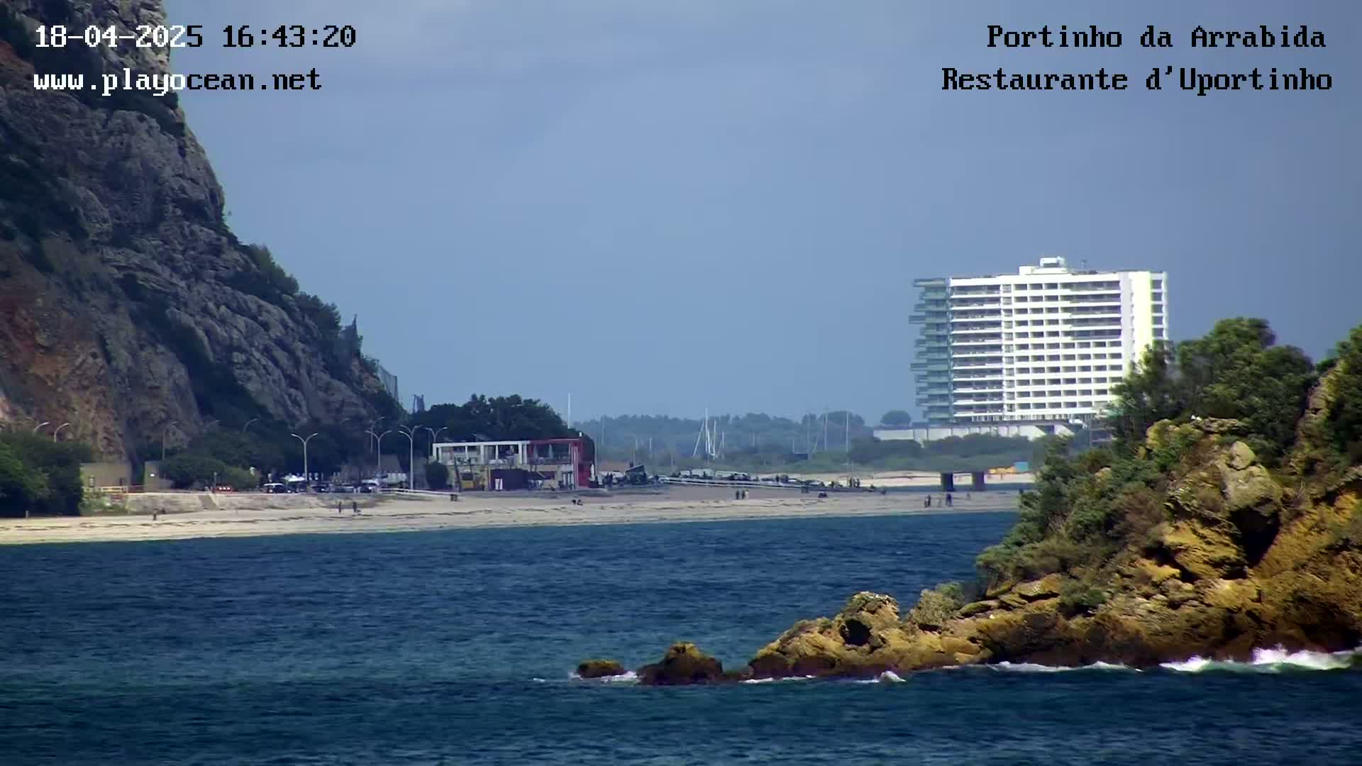 A sandy beach with a few people on it stretches between a rocky cliff and a large building, with a rocky islet in the foreground and calm, blue water.